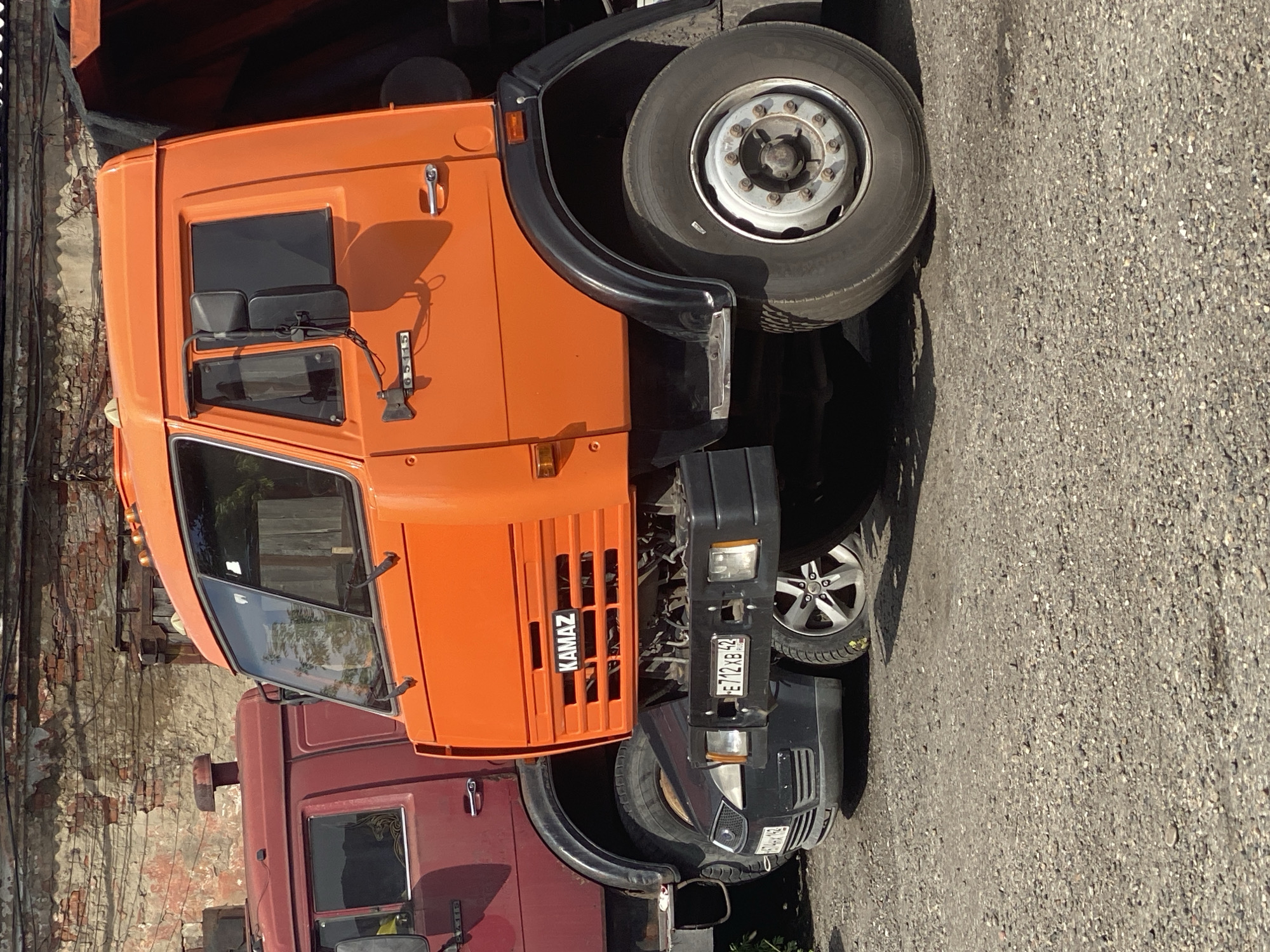 The image shows the side view of two large trucks parked on a gravel surface in Novokuznetsk, Russia. The truck in the foreground is bright orange and appears to be a dump truck, with the brand name "KAMAZ" visible on its grille. It has large wheels and a robust chassis. Partially visible behind the orange truck is a dark red truck. In between the two trucks, a black sedan is parked. The background features a dilapidated brick building with visible power lines and weathered walls. The scene appears to be an outdoor industrial or service area.
