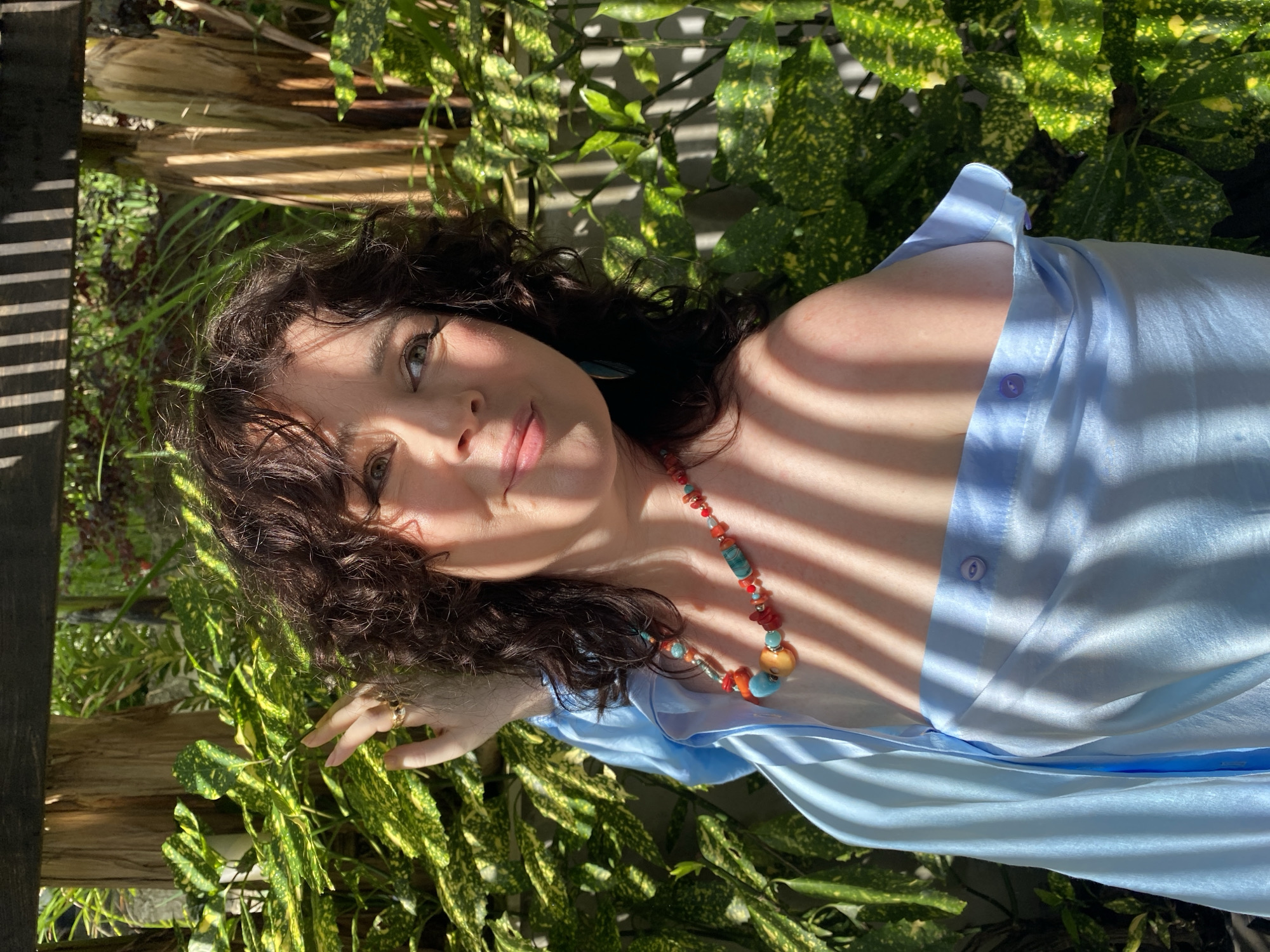 A medium shot shows a woman with curly brown hair and fair skin posing outdoors, with a blurred background of lush green foliage and wooden structures. She is wearing a light blue, off-the-shoulder silk top adorned with a colorful beaded necklace featuring orange, red, turquoise, and blue stones. Her right hand is raised, with fingers slightly splayed, near her head, and she wears a gold ring on her index finger. Sunlight casts striped shadows across her face and shoulders. The setting appears to be a garden or courtyard, possibly in Sochi, Russia, given the vibrant greenery and implied warm climate.