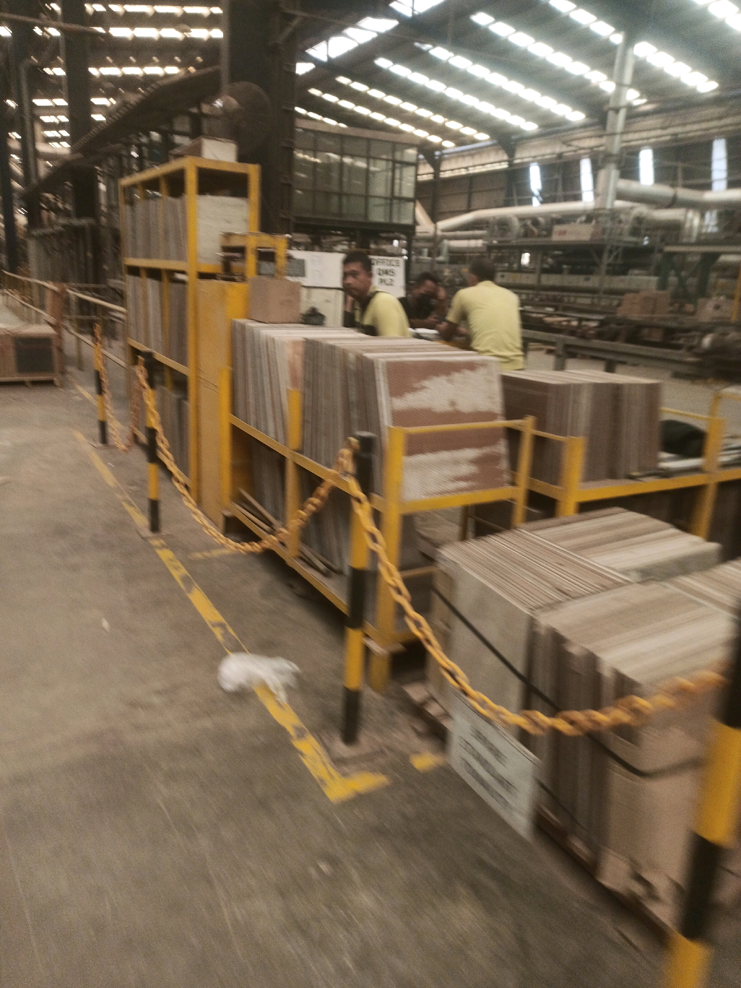 The image shows an indoor industrial setting, likely a factory or warehouse, in Lebaniwaras, Indonesia. The scene is characterized by its large scale and the presence of industrial equipment and materials.
In the foreground, stacks of rectangular objects, possibly tiles or construction materials, are neatly arranged on shelving units. These units are painted in a bright yellow color, which contrasts with the muted tones of the materials and the surrounding environment. Yellow and black striped bollards with yellow chains are present, indicating a designated walkway or safety zone.
In the mid-ground, two men are visible. One man, wearing a yellow shirt, is seated and appears to be looking towards something out of frame. Another man, also in a yellow shirt, is standing and looking away from the camera. The presence of these individuals suggests that the location is operational.
The background reveals the extensive structure of the building, with a high, sloped roof featuring numerous skylights or translucent panels that allow natural light to filter in. Various pipes, conduits, and industrial machinery are visible, indicating a complex manufacturing or processing facility. The overall impression is one of a functional, working industrial space.