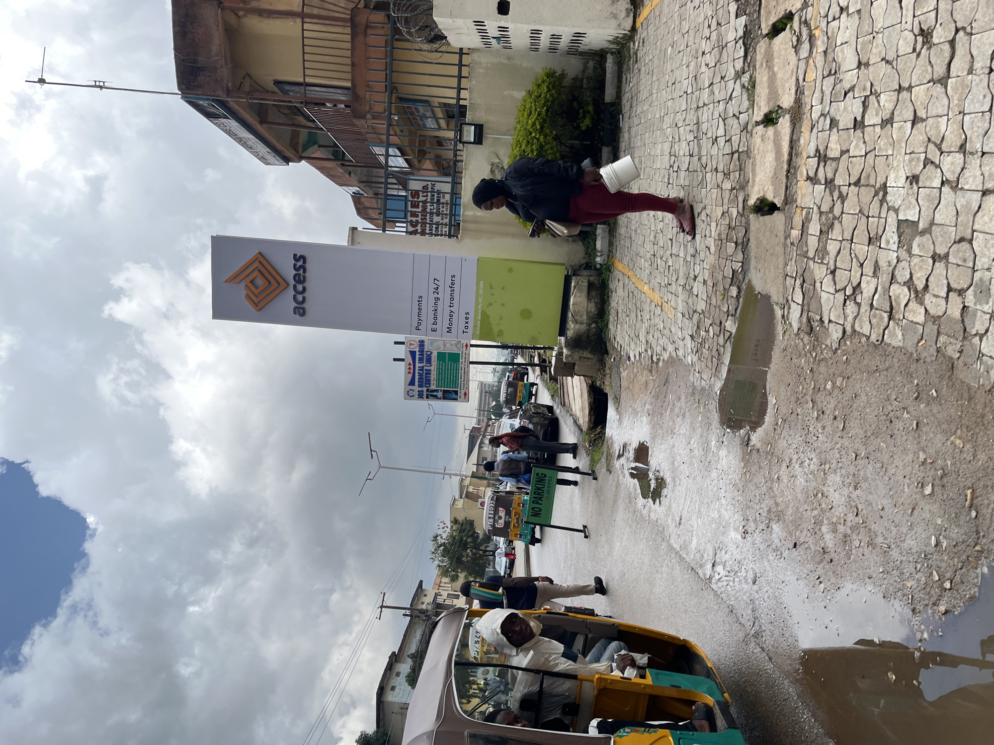 This image captures a street scene in Jos, Nigeria, on a cloudy day, with evidence of recent rain creating puddles on the cobbled pavement and road. A woman in a black jacket and red trousers is walking, carrying a white bucket and a bag. She appears focused on her phone, which she holds in her left hand.
Several vehicles are visible on the street, including a yellow auto-rickshaw in the foreground with a driver wearing a white hooded garment and passengers. In the background, other vehicles and people are seen, suggesting a typical urban environment.
A prominent "Access" bank sign stands tall, displaying services like "Payments," "E-banking 24/7," "Money transfers," and "Taxes." Next to it, a "NO PARKING" sign is also visible. Other signs in the background include "JOS MEDICAL IMAGING CENTRE (JMIC)" and "ACFES NDISE NIC.LTD. LABORATORY, SCIENTIFIC." The overall impression is a busy street with commercial activity, common in many Nigerian cities. The weather appears overcast, contributing to the dampness of the surroundings.