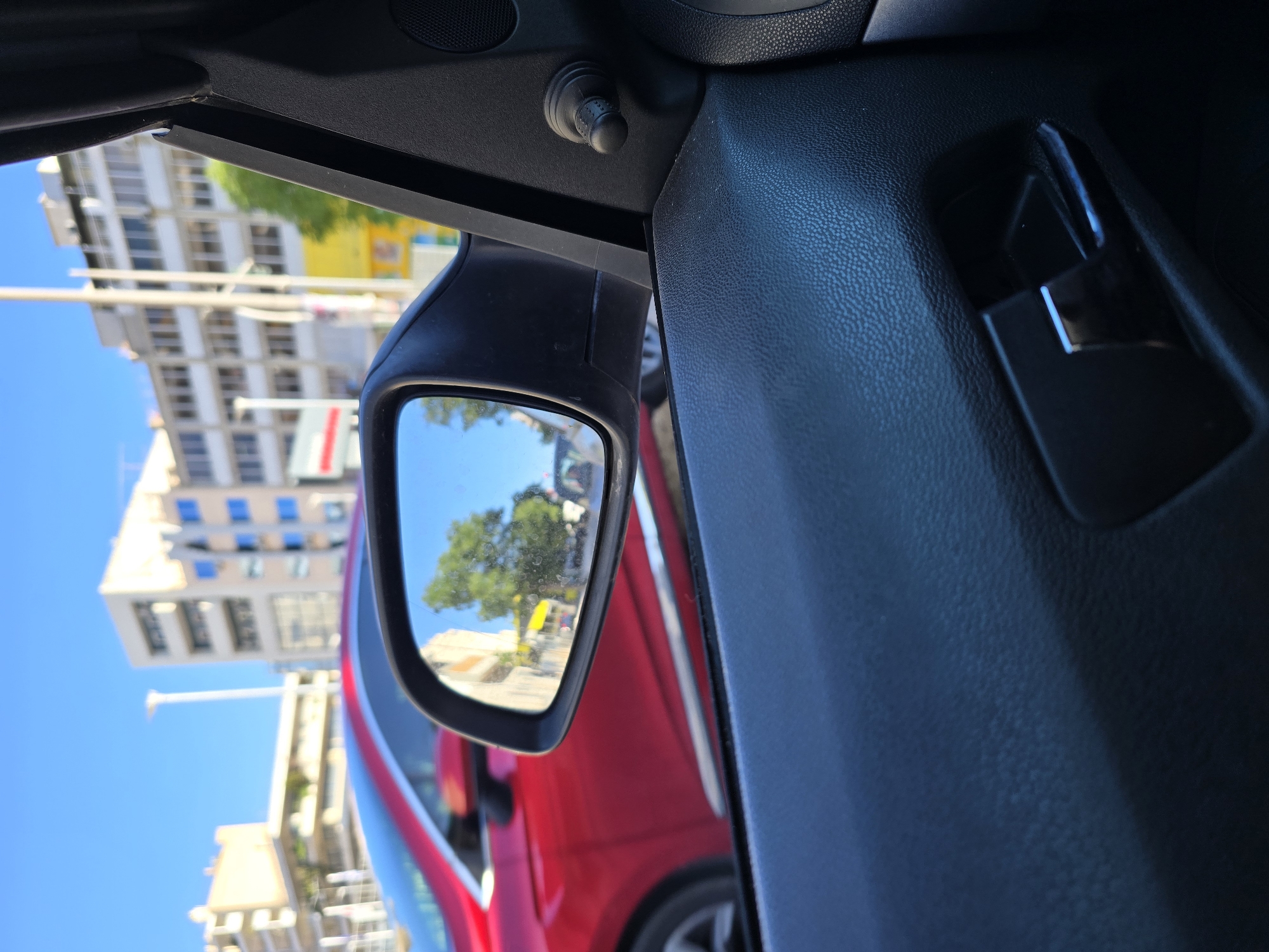 This image captures a view from inside a car, looking out the side window towards a street scene in Athens, Greece. The focus is on the car's side mirror, which reflects a sunny day with buildings and trees. A bright red car is visible in the foreground, to the left of the mirror, with its side profile in view. The interior of the car, including the door panel and a handle, is also visible. The background shows urban architecture with buildings of varying heights and a clear blue sky. The scene appears to be taken during daylight, possibly late afternoon, given the lighting and shadows.