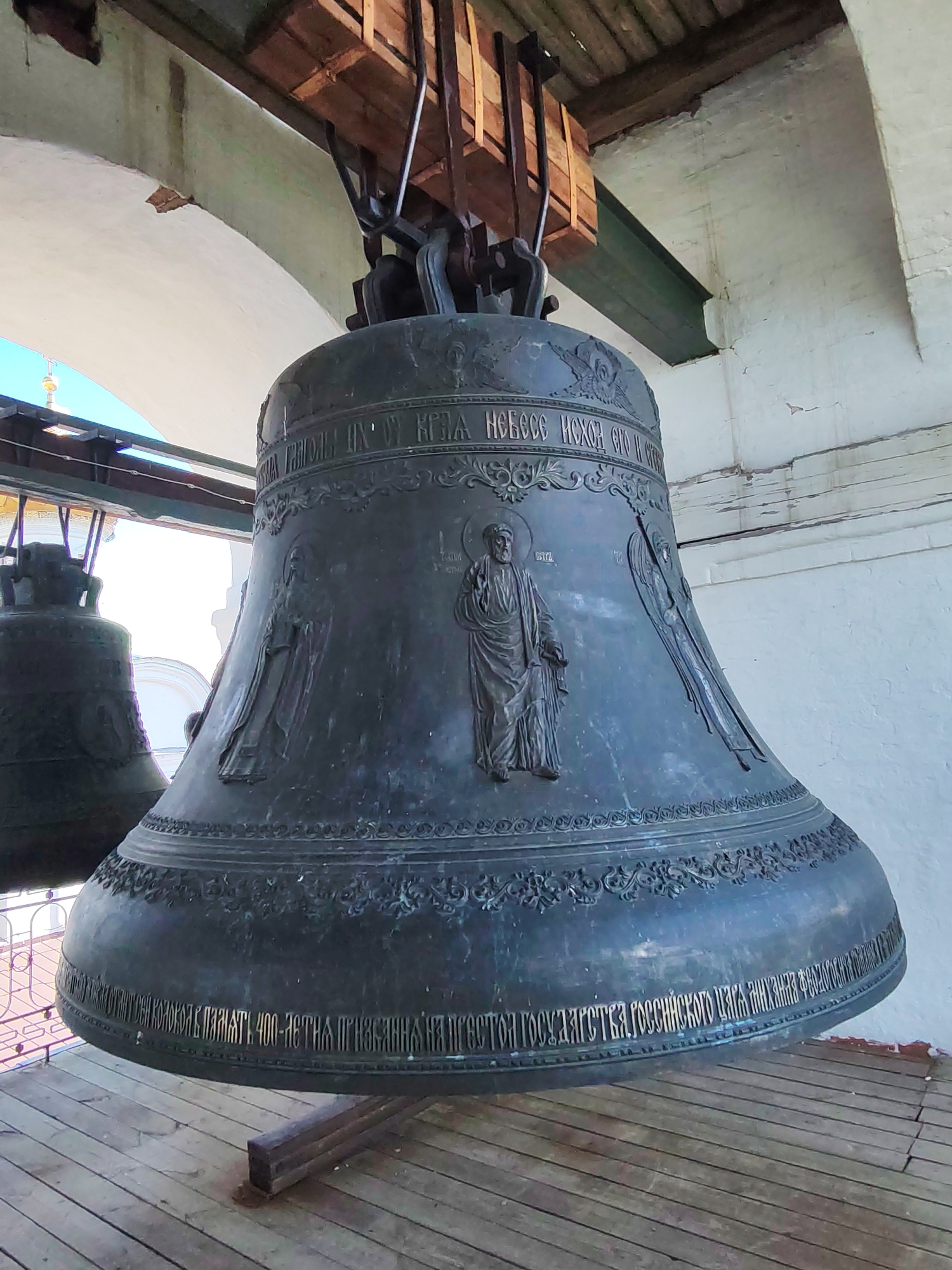 This image shows a close-up view of a large bronze bell, likely the Tsar Bell, in Kostroma, Russia. The bell is ornately decorated with religious imagery and Cyrillic inscriptions. It is suspended from a wooden structure with visible metal supports, suggesting it is part of a bell tower or a similar church structure. The bell is dark metallic in color and shows signs of age and patination. In the background, a second, similar bell is partially visible, also dark in color. The bells are situated on a wooden platform, supported by beams, with wooden planks forming the floor. The architecture of the surrounding structure is visible, with a white, textured wall and arched openings that reveal a bright, outdoor environment with a clear sky and architectural elements of other buildings, including a spire-like structure. The lighting suggests it is daytime, possibly sunny.