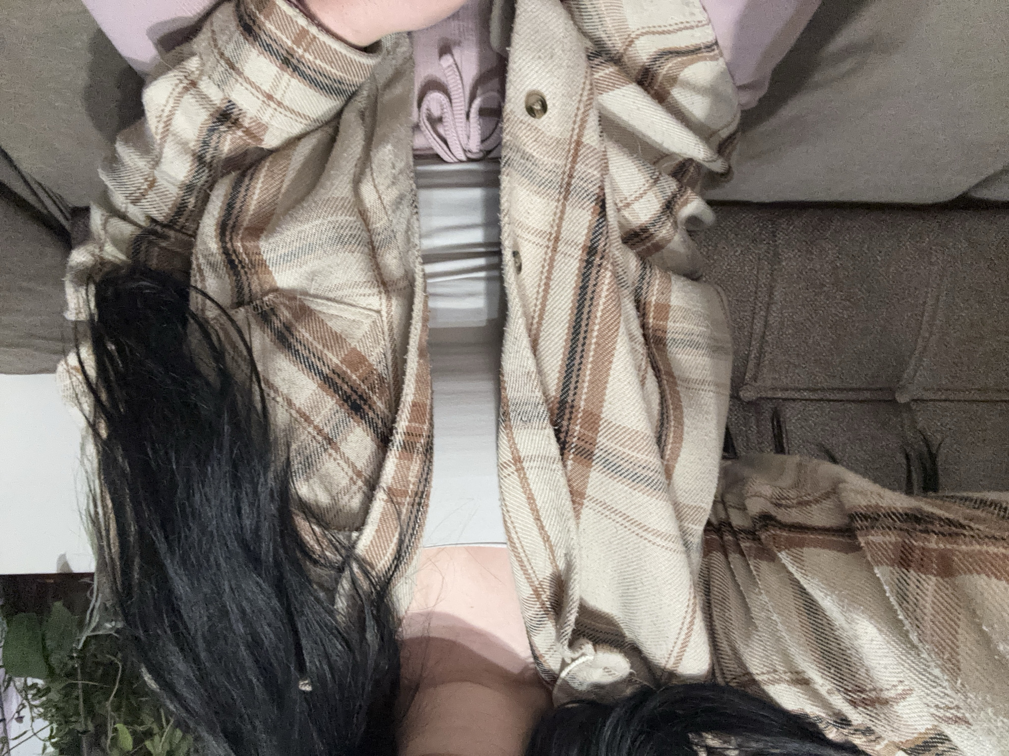 A close-up shot, taken from an overhead perspective, depicts a person sitting on a grey sofa. The person is wearing a pink hooded top, a white top with grey stripes, and a tan and brown plaid shirt. Long, black hair is visible on the left and lower right of the frame. The shot is taken indoors. It is impossible to place the image in the city of Asenovgrad, Bulgaria based on provided image information.