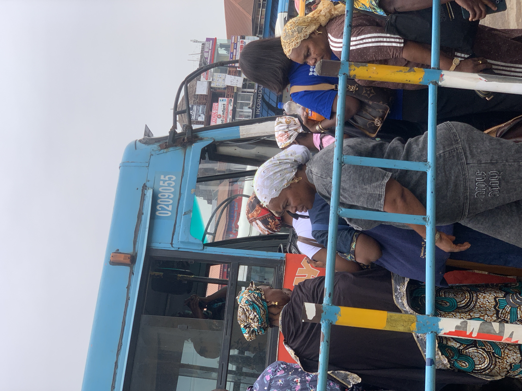 A group of people are waiting next to a blue bus in Ikorodu, Nigeria. The bus is numbered "0209055". The people are pressed closely together, holding onto the blue metal bars of the bus. Some of them are wearing traditional head coverings. A series of storefronts and buildings are visible in the background, with multiple signs posted. The sky is overcast and gray.