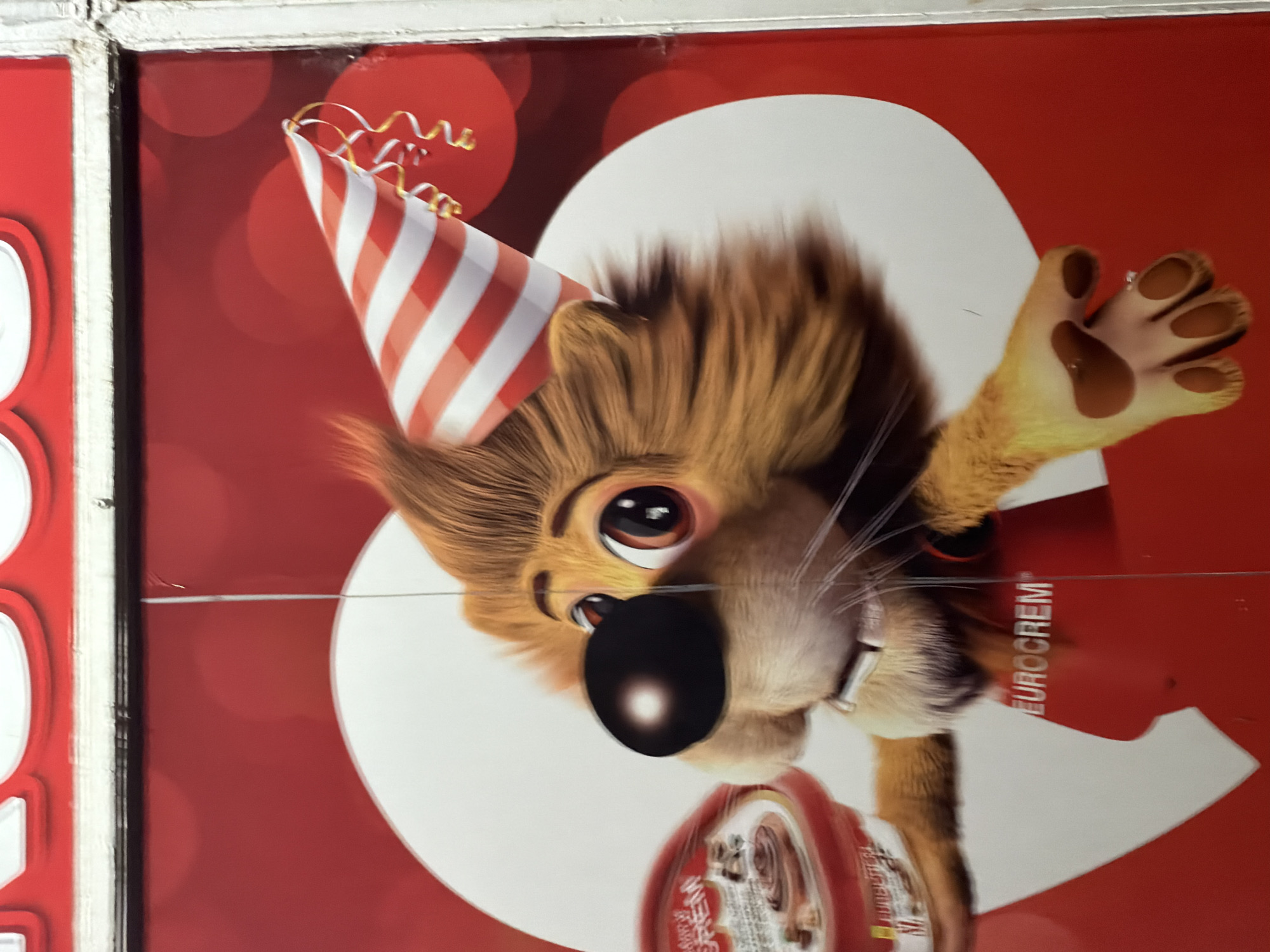 This media file displays a promotional advertisement featuring a cheerful, cartoon-style lion mascot. The lion wears a red and white striped party hat with yellow streamers, suggesting a celebratory theme. It has large, expressive eyes and a slight smile, with one paw raised as if waving. In its other paw, the lion holds a red, cylindrical product container, likely a jar or can, though the text on it is mostly illegible.
The background is a vibrant red with soft, blurred circles in a lighter red tone, creating a festive atmosphere. A large, abstract white shape is positioned behind the lion's head. The most prominent visible text is "EUROCREM," oriented vertically along the right side of the image.
The image appears to be a physical display, such as a poster or sign, indicated by a faint white frame on the left and top edges, and some minor horizontal and vertical lines across the surface, which might be creases, scratches, or wires. The overall impression conveyed is one of fun and happiness, typical of product advertising. This display is located in Podgorica, Montenegro. The entire image is captured with a 90-degree counter-clockwise rotation from its likely intended upright orientation.