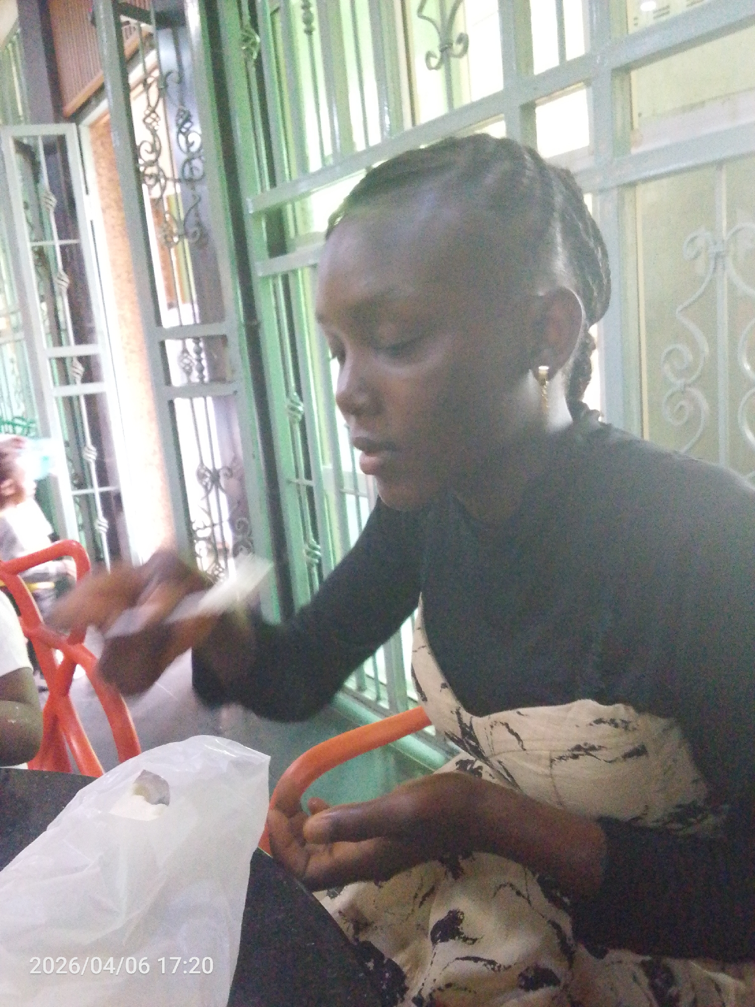 A woman with braided hair is seated at a table indoors. She is wearing a black long-sleeved shirt and a patterned dress. In front of her is a white plastic bag. She is holding something in her hands. Other people are partially visible in the background, seated at an orange chair. A window with ornate metal bars is behind her. The timestamp "2026/04/06 17:20" is visible at the bottom of the image. The city of Lokoja, Nigeria, is not explicitly identified as the location in the image.