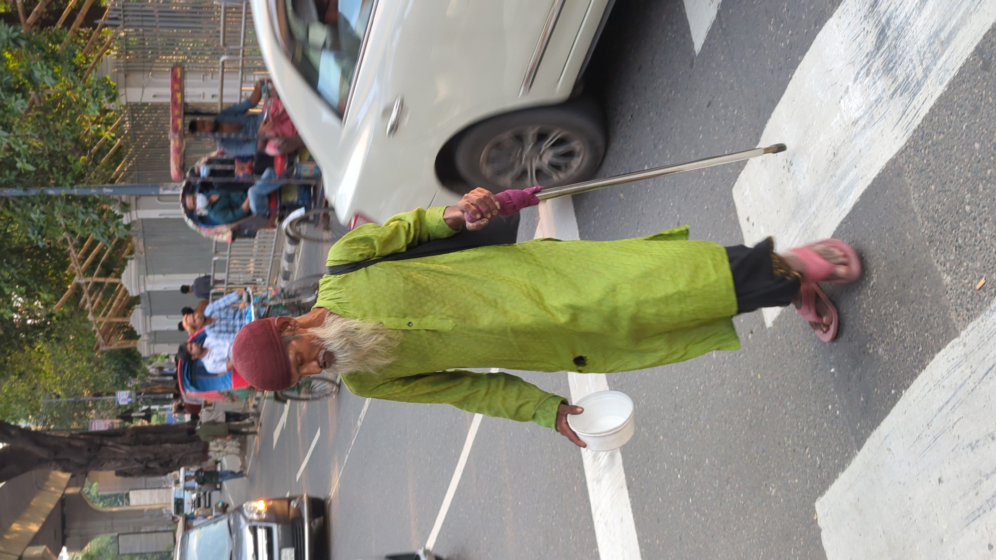 A street in Dhaka, Bangladesh features a pedestrian crossing with a blind man dressed in green clothing. He's wearing a red cap and sporting a long white beard. He holds a white bowl in one hand and uses a cane for guidance. A white car and motorized rickshaws are visible in the background.