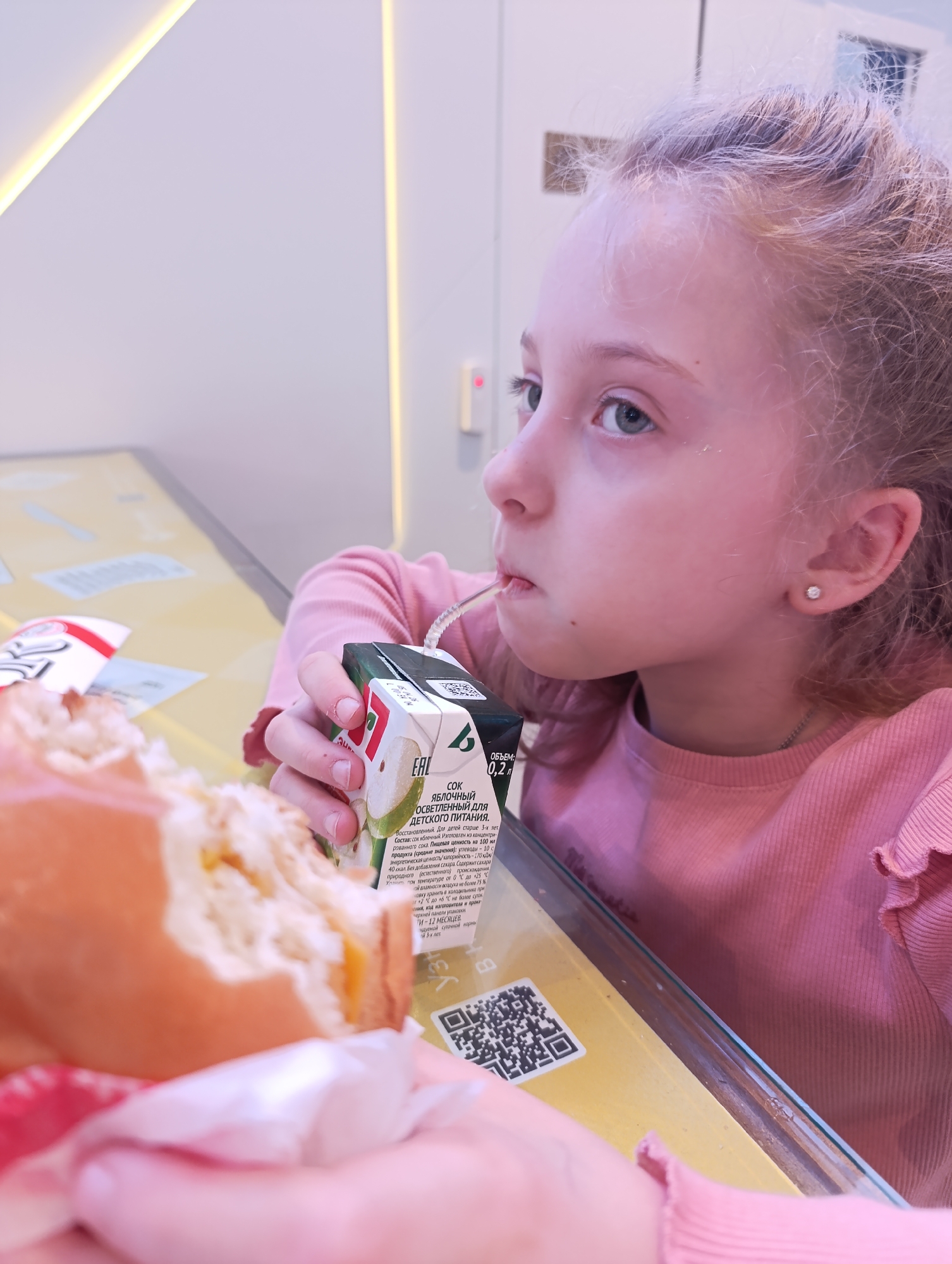 A young, fair-skinned girl with light blonde hair is drinking juice with a straw at a table. She is wearing a pink long-sleeved shirt. The juice box is labeled in Russian as "COK YABLOCHNYI OSVETLENNYI DLYA DETSKOGO PITANIYA" which translates to "Apple Juice Lightened for Baby Food". Next to her is a partially eaten sandwich in a wrapper. The table has a yellow surface under a glass top. A QR code is visible on the table. The location appears to be indoors, possibly a cafe or restaurant. The city of Moscow, Russia, is not indicated in the image.