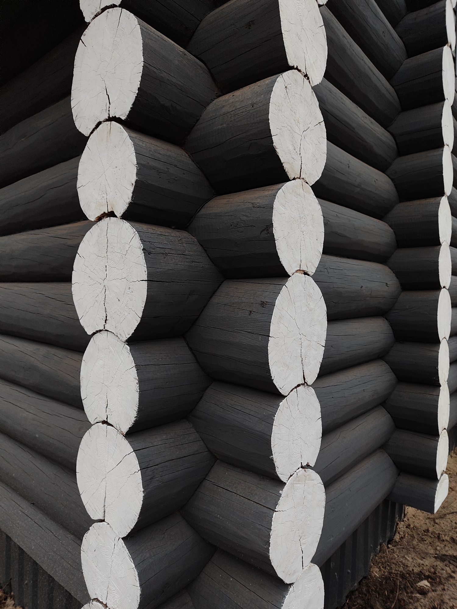 The image shows a close-up of the corner of a log cabin made of dark, possibly charcoal-colored, logs with white-painted ends. The logs are stacked horizontally, interlocking at the corner. The ground near the base of the cabin is dirt. The location is Unknown, Unknown.