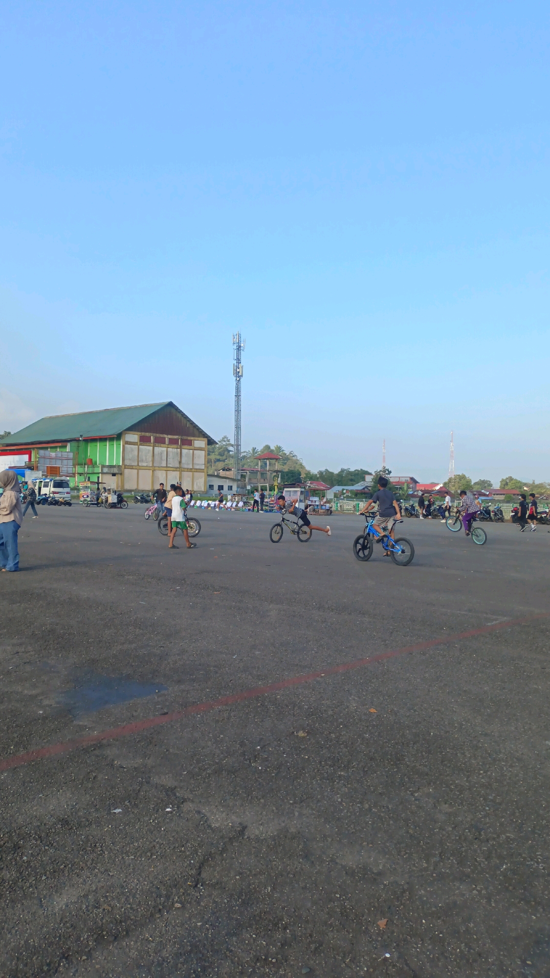 In Padang Panjang, Indonesia, this image captures an outdoor scene with a clear, light blue sky. A large, flat, paved area, likely a parking lot or open space, dominates the foreground. Several children are actively engaged in riding bicycles, some in motion and some stationary. A prominent building with a green roof and white walls stands in the background, possibly a community center or stadium. A tall cellular tower is visible in the distance, along with other smaller buildings and trees. The ground is asphalt, with some scuffs, cracks, and a faint red line marking. A few vehicles, including what appear to be cars and motorcycles, are parked or moving in the middle ground. The overall atmosphere suggests a leisurely daytime activity.