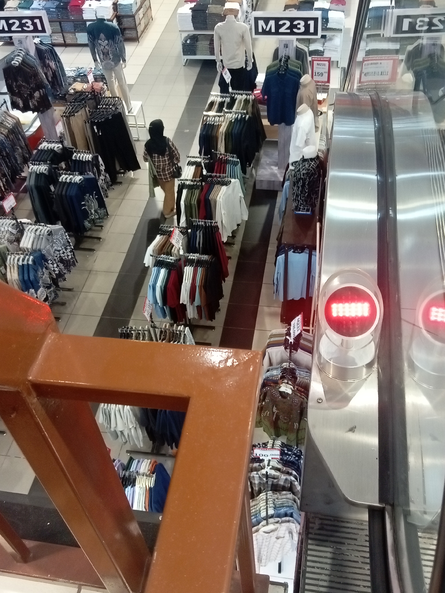 The image captures a high-angle view of a clothing retail store, likely in Batam, Indonesia, given the signage "M231" which is a common department store chain in Indonesia. The store appears to be well-stocked with men's clothing, displayed on racks and shelves. In the foreground, a brown railing overlooks the store. To the right, a section of a silver escalator with red digital displays is visible.
In the middle ground, a woman wearing a black hijab and a plaid shirt is walking through the store. Several mannequins are dressed in clothing and positioned throughout the aisles, showcasing various garments. Racks of shirts, trousers, and other apparel are neatly arranged. The store's flooring is a tiled pattern of light and dark grey squares. Signs with prices and product codes, such as "M231" and "159,000", are displayed. The lighting suggests an indoor environment, typical of a department store.
