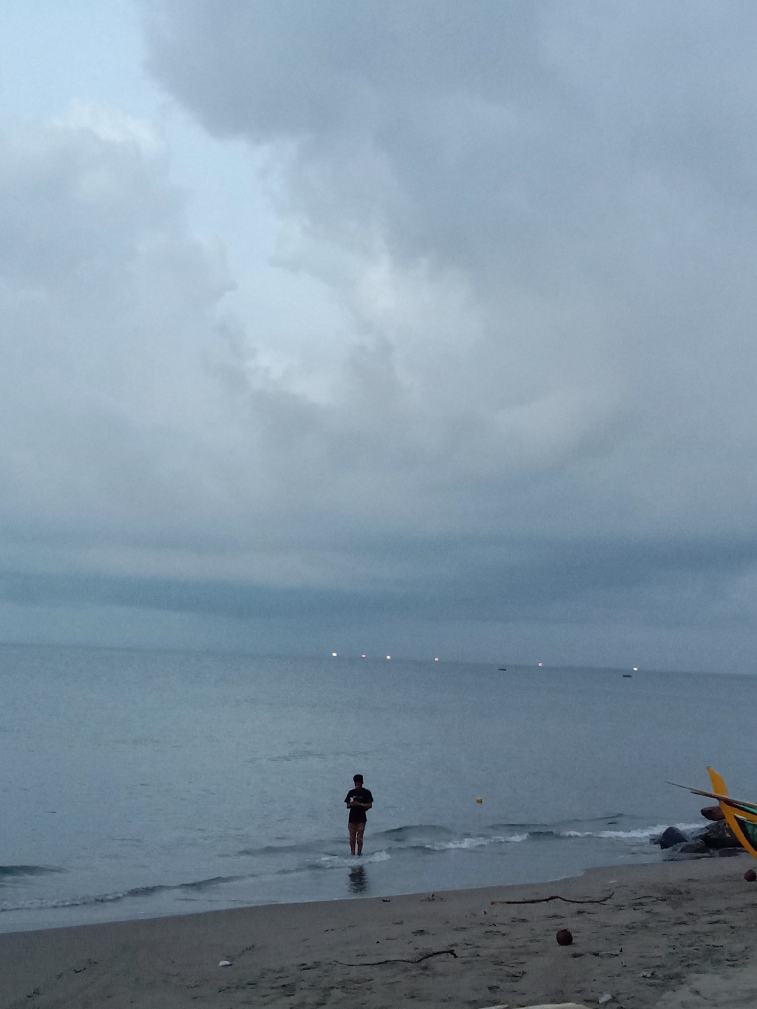 The image depicts a coastal scene in Lhokseumawe, Indonesia. Overcast skies dominate the upper portion of the frame, casting a somber light over the landscape. A calm body of water stretches horizontally across the middle ground, with several distant lights visible on the horizon, likely from boats or coastal structures.
In the foreground, a person stands ankle-deep in the gentle waves at the edge of a sandy beach. The individual is wearing a dark t-shirt and shorts. To the right, part of a traditional Indonesian boat is visible, along with some rocks. Debris, including a coconut, is scattered on the sand.