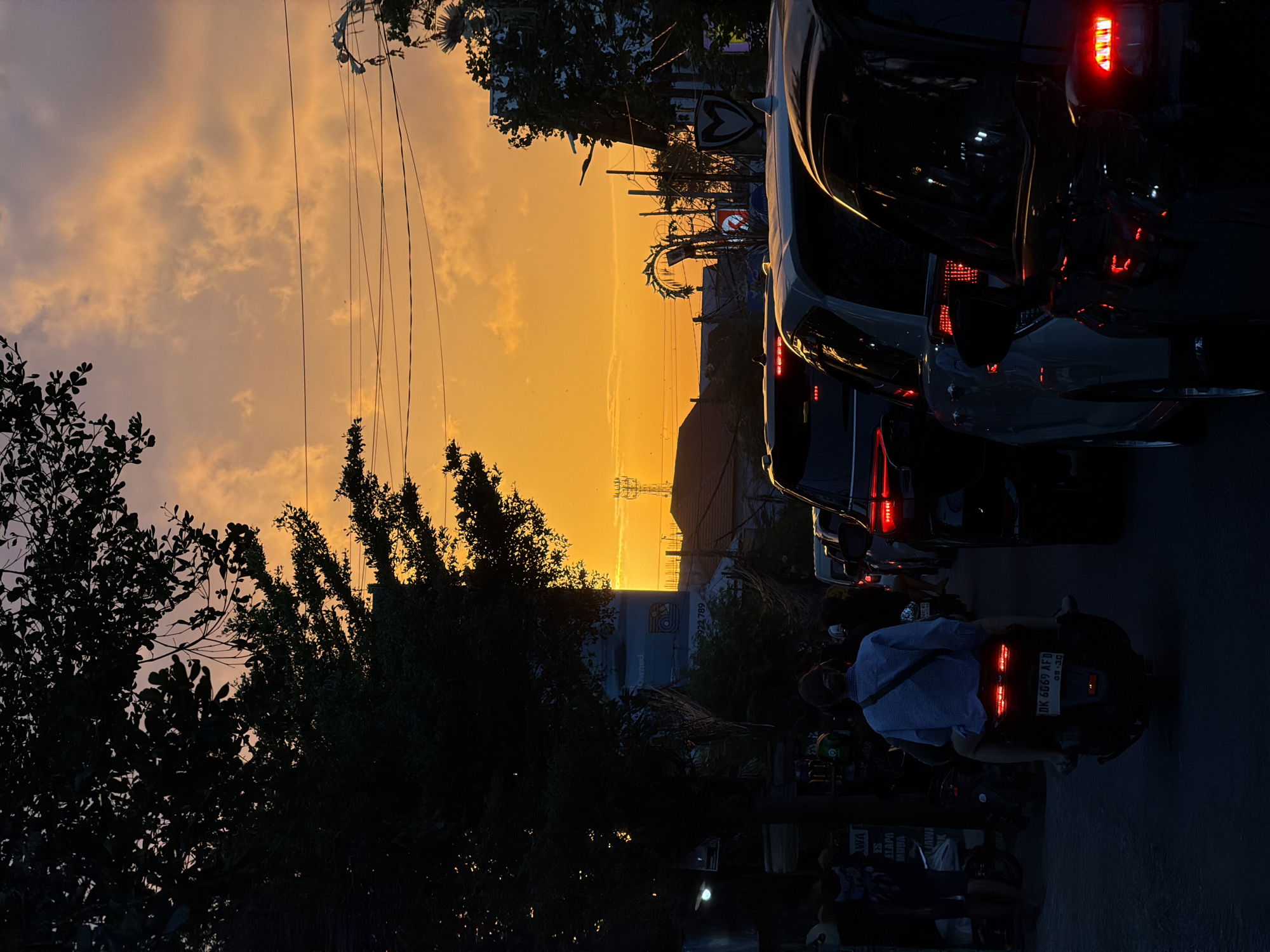 This is a photograph of a street scene in North Kuta, Indonesia, during a sunset. The sky is a vibrant orange and yellow, with scattered clouds. In the foreground, several cars are stopped in traffic, their brake lights illuminated. A motorcycle is also present, carrying two people. The scene is silhouetted against the bright sky, with trees and buildings visible in the background. The image captures the atmosphere of a typical evening commute in this Indonesian city.