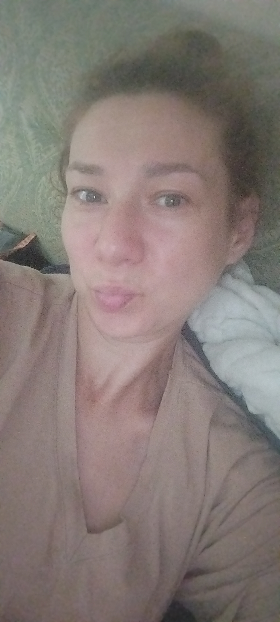 A medium shot, taken from a slightly elevated perspective, shows a woman lying down in what appears to be a bed. She has her hair pulled up and is wearing a tan v-neck shirt. Her face is close to the camera, and she is making a kissing face, with her lips pursed. Her eyes are open and looking towards the camera. There is a white blanket or pillow behind her right side, and a green patterned surface to her left. The lighting is soft and diffused. There is no specific detail that allows for the identification of the city of Mariupol, Ukraine.