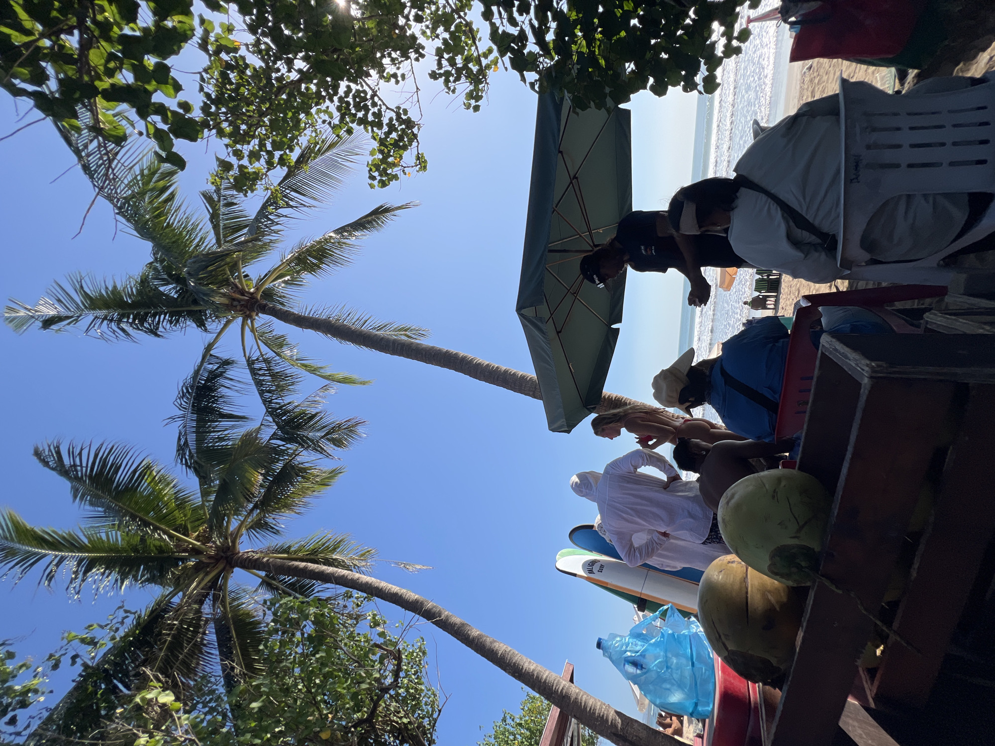 In Kuta, Indonesia, on a bright, sunny day, a group of people are gathered on a sandy beach near palm trees. In the foreground, a wooden crate holds several coconuts. Behind the crate, several surfboards stand upright, leaning against each other. A person in a white hooded outfit stands near the surfboards, facing away from the camera. To their right, a woman in a bikini stands with her back to the viewer. Further back, under a large green umbrella, a man in a black t-shirt stands. Several individuals are seated in chairs facing the ocean, which features moderate waves. The overall scene suggests a relaxed beach environment with activities like surfing and enjoying the outdoors.