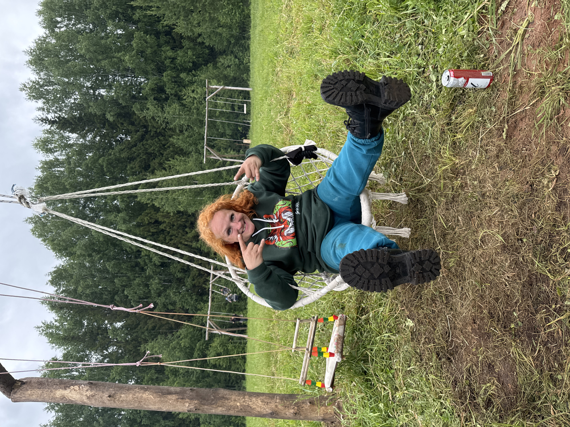 An adult individual, presumably female, with bright red, curly hair, is seated in a white, woven hanging swing chair in an outdoor grassy field in Bol'shaya Kivara, Russia. She is wearing a dark green hooded sweatshirt featuring a colorful graphic of a stylized fox head with antlers, teal blue athletic pants, and black, lug-soled boots. Her right hand is raised, with the index finger pointed towards her face and the thumb and pinky extended in a gesture. She is smiling directly at the viewer.
To her immediate left, a smaller, unoccupied wooden swing with colorful red, yellow, and green accents hangs from a thick, rustic wooden post. In the middle distance, another wooden swing set is visible, with several indistinct figures, possibly children, using the swings. A dense line of green trees forms the background. The sky is overcast and grey. On the muddy, grass-covered ground to the lower right of the seated individual, a red and white aluminum beverage can (appears to be a Baltika 9 beer can) lies on its side.