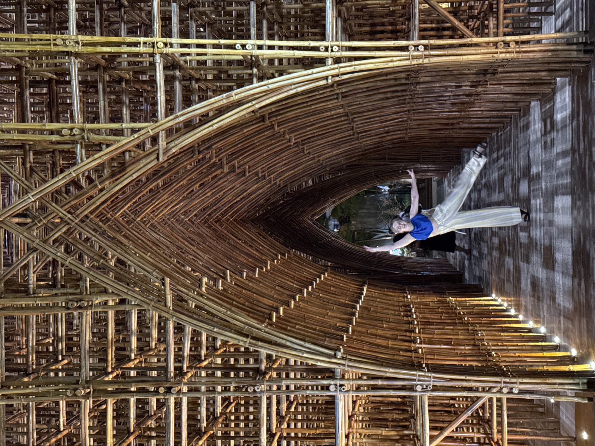 This vertical image features a young woman posing gracefully within an intricately designed architectural structure made entirely of bamboo. The woman, smiling and with her left arm raised, is wearing a blue crop top and light-colored, striped wide-leg pants. She is positioned on a dark wooden floor, within what appears to be a large, arching bamboo tunnel or walkway.
The bamboo structure itself is highly detailed and complex, showcasing various patterns of interwoven and stacked bamboo poles. It forms a deep, elongated arch that leads into a darker background, which hints at greenery, suggesting an outdoor or semi-outdoor setting beyond the immediate tunnel. The warm, yellowish glow from small, embedded lights along the inner curves of the bamboo structure illuminates the scene, casting soft highlights and shadows, and indicating that the photo was taken during the evening or night. The intricate craftsmanship of the bamboo, varying in shades of brown and tan, creates a visually rich and immersive environment. The woman's dynamic pose adds a sense of movement and artistry to the unique setting, characteristic of some modern designs found in Phú Quốc, Vietnam. No visible text is present.