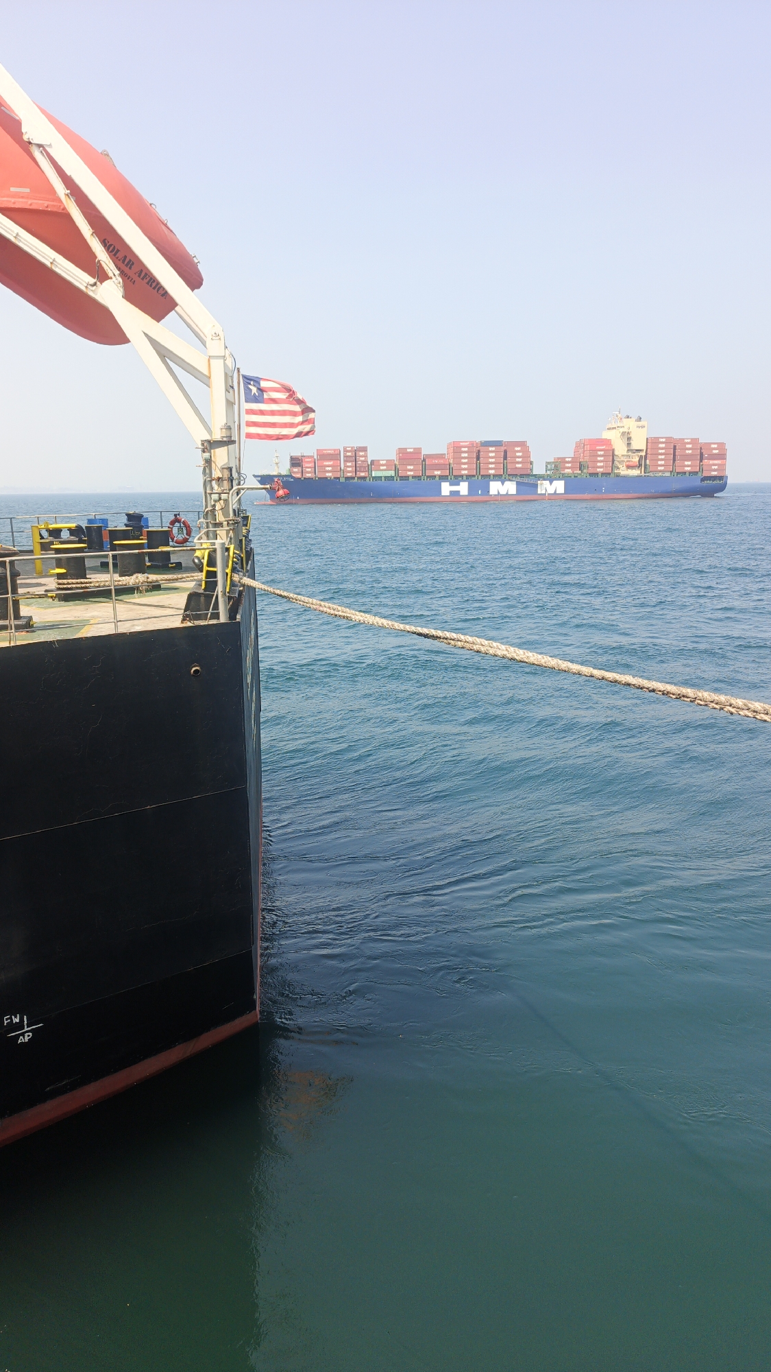 This is a photograph taken from the deck of a ship looking out towards another ship. The ship in the foreground is black and has a red waterline. A life raft, also red, is visible on the left side of the frame, with "SOLAR AFRICA" printed on it. A Liberian flag is also visible on the front ship. The ship in the background is a large container ship with "HMM" printed on its side, carrying numerous red shipping containers. The ships are at sea, with calm blue water. The sky is clear and bright. The scene appears to be a maritime setting, possibly in the waters off Singapore, given the prevalence of shipping traffic in the region.
