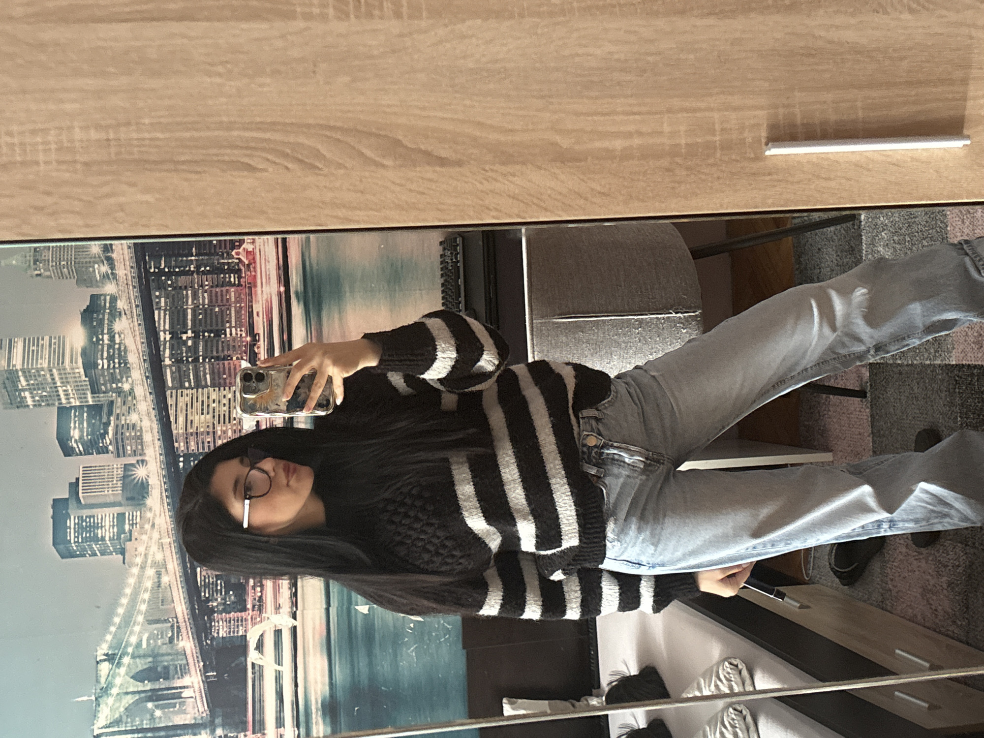 A woman is taking a mirror selfie. She is wearing glasses, a black and white striped sweater, and light blue jeans. The reflection in the mirror includes a cityscape backdrop and a room with a wooden dresser. There is no indication of a location in Sofia, Bulgaria.