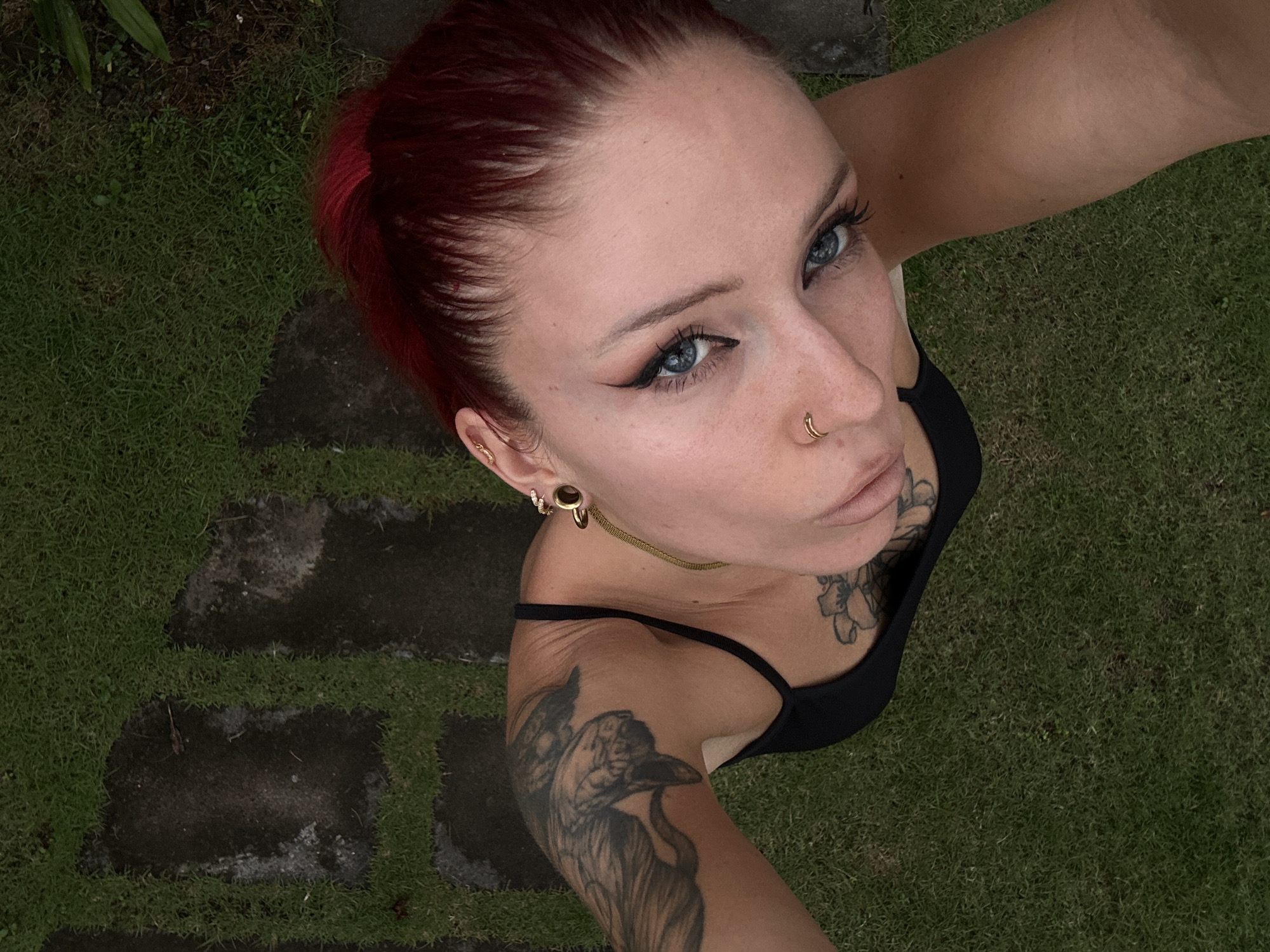 A high-angle, close-up shot shows a fair-skinned person with bright blue eyes and winged eyeliner, looking upwards. Their auburn hair is pulled back. They are wearing a black tank top, a gold hoop earring, and a gold nose ring. Tattoos are visible on their arm and chest. The person is outdoors, with a background of green grass and dark, wet paving stones. This scene is located in North Kuta, Indonesia.