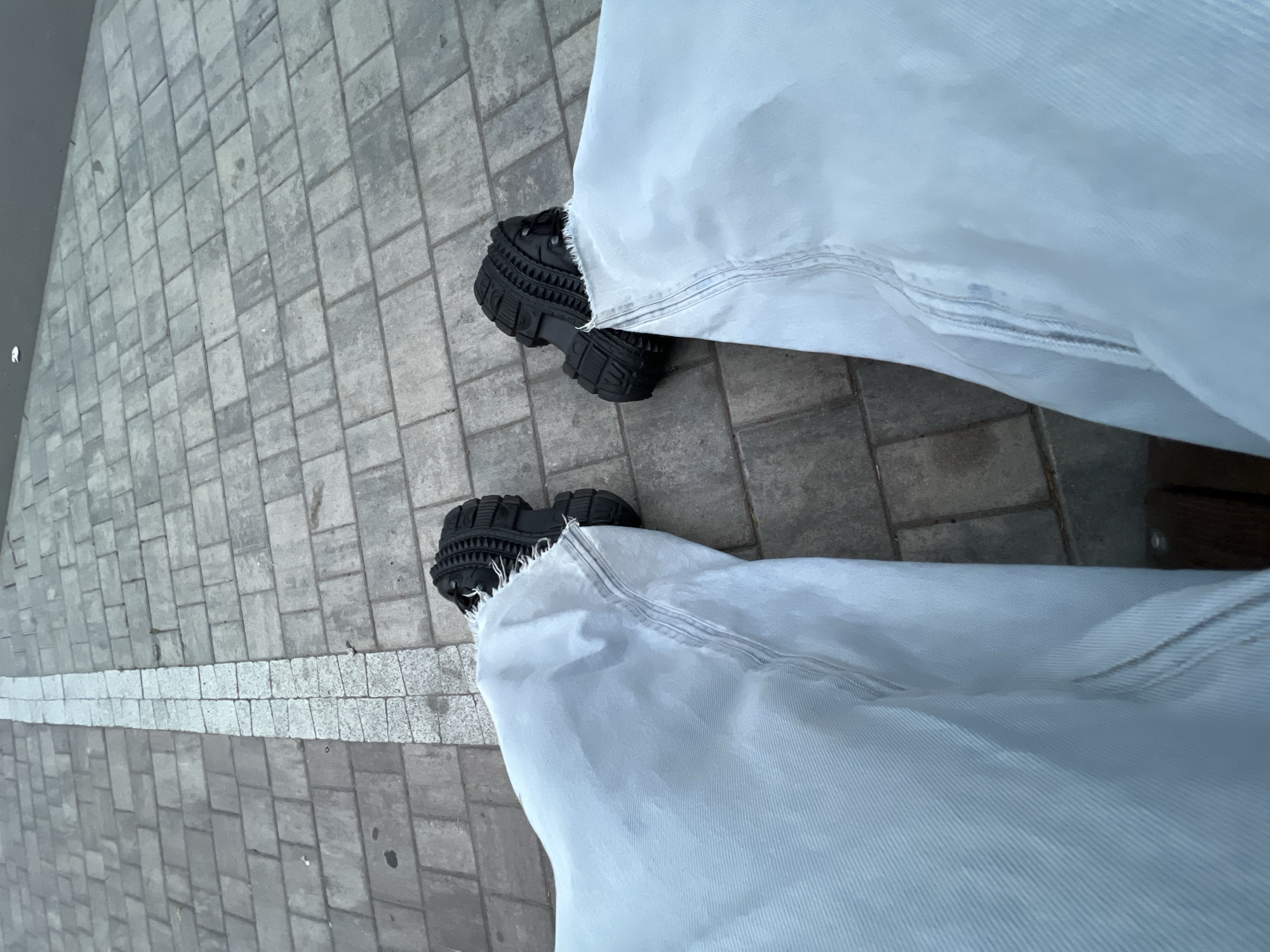 The image depicts a first-person, downward-looking view of a person's lower legs and feet on a paved outdoor surface. The individual wears light blue or off-white wide-leg pants with frayed cuffs and black, chunky-soled shoes with aggressive tread patterns. The ground is paved with rectangular grey concrete blocks. A prominent, straight line of smaller, lighter-colored rectangular granite pavers runs centrally from the bottom center into the distance, separating the grey paving. To the right, in the upper background, the paved area transitions to a darker, smoother surface, likely asphalt. This specific paving pattern, with its distinctive light granite line set within grey pavers, is characteristic of the **Griboyedov Canal embankment in Saint Petersburg, Russia, particularly in the vicinity of the Church of the Savior on Spilled Blood**.