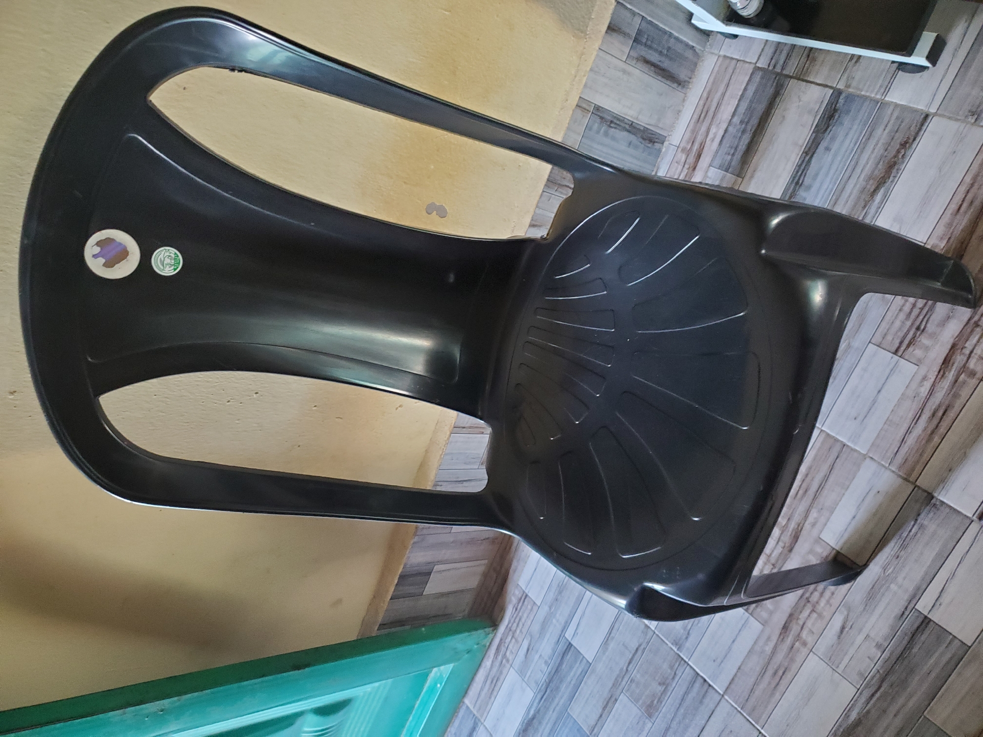 A black plastic chair is shown in a room. The chair has a molded seat with a radiating pattern and a curved back. There are two stickers on the back of the chair, one circular and white with some purple markings, and another smaller green and white circular sticker below it. The chair is positioned on a tiled floor with a wood-plank pattern. To the left of the chair is a partial view of a green door frame. The wall behind the chair is a light yellow color, with some small water stains visible. The scene appears to be indoors in Jauro Yinu, Nigeria.
