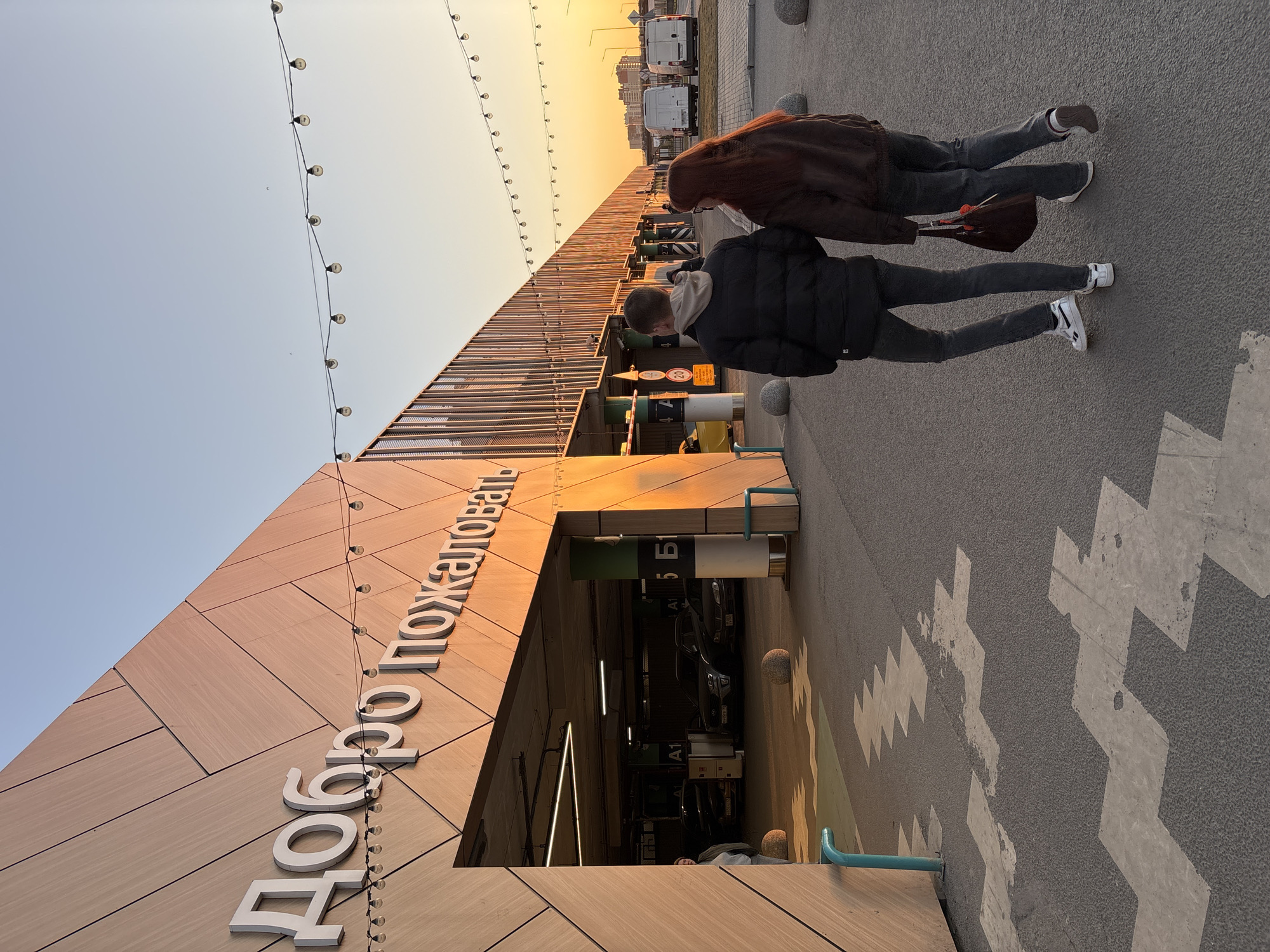 The image captures an exterior scene in what appears to be Kudrovo, Russia, as implied by the Cyrillic inscription "Добро пожаловать" (Welcome) on the building. The photo is taken at ground level, looking up and slightly to the left. Two people are walking in the direction of the camera, one slightly ahead of the other. Both are wearing dark clothing; the forward individual has dark pants and white shoes, while the following one has gray pants and gray shoes. The building has a modern architectural design. The entrance to a parking garage is visible. A string of lights is suspended above the structure. The sky has a gradient of colors, suggesting either sunrise or sunset. The pavement has a white chevron pattern.