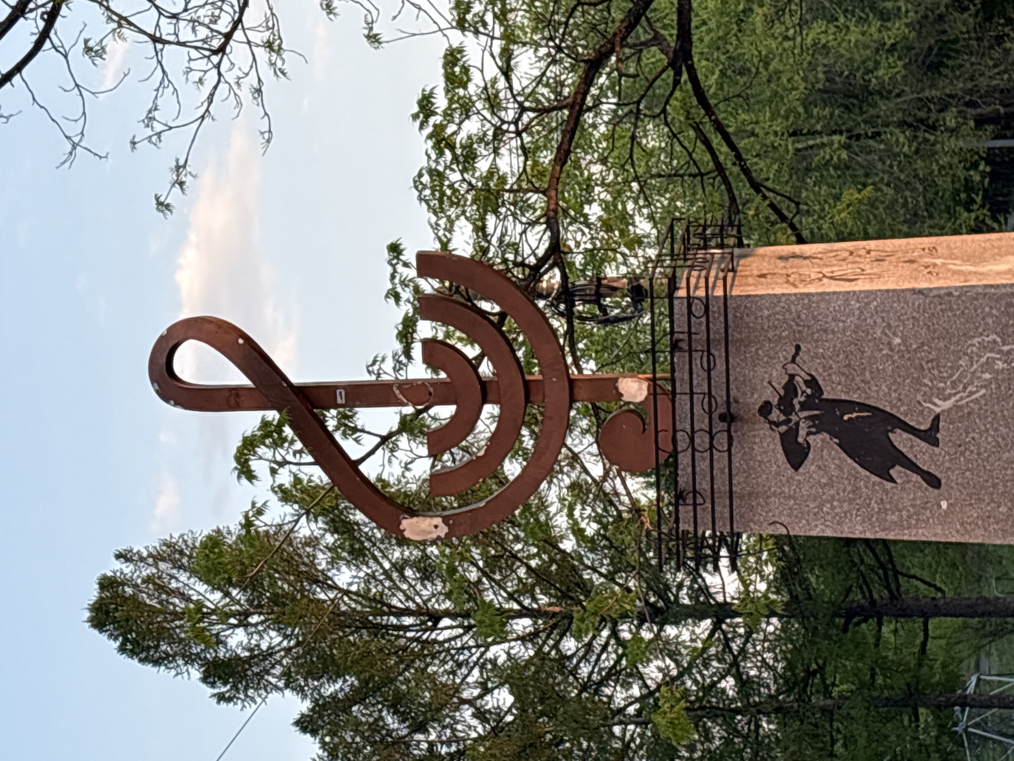 An outdoor monument in Birobidzhan, Russia, features a rusty metal sculpture resembling a musical notation symbol attached to a concrete pillar with an illustration of a man playing the violin. The monument is framed by trees and a section of the sky. A metal fence with a design of musical bars is behind the concrete pillar.