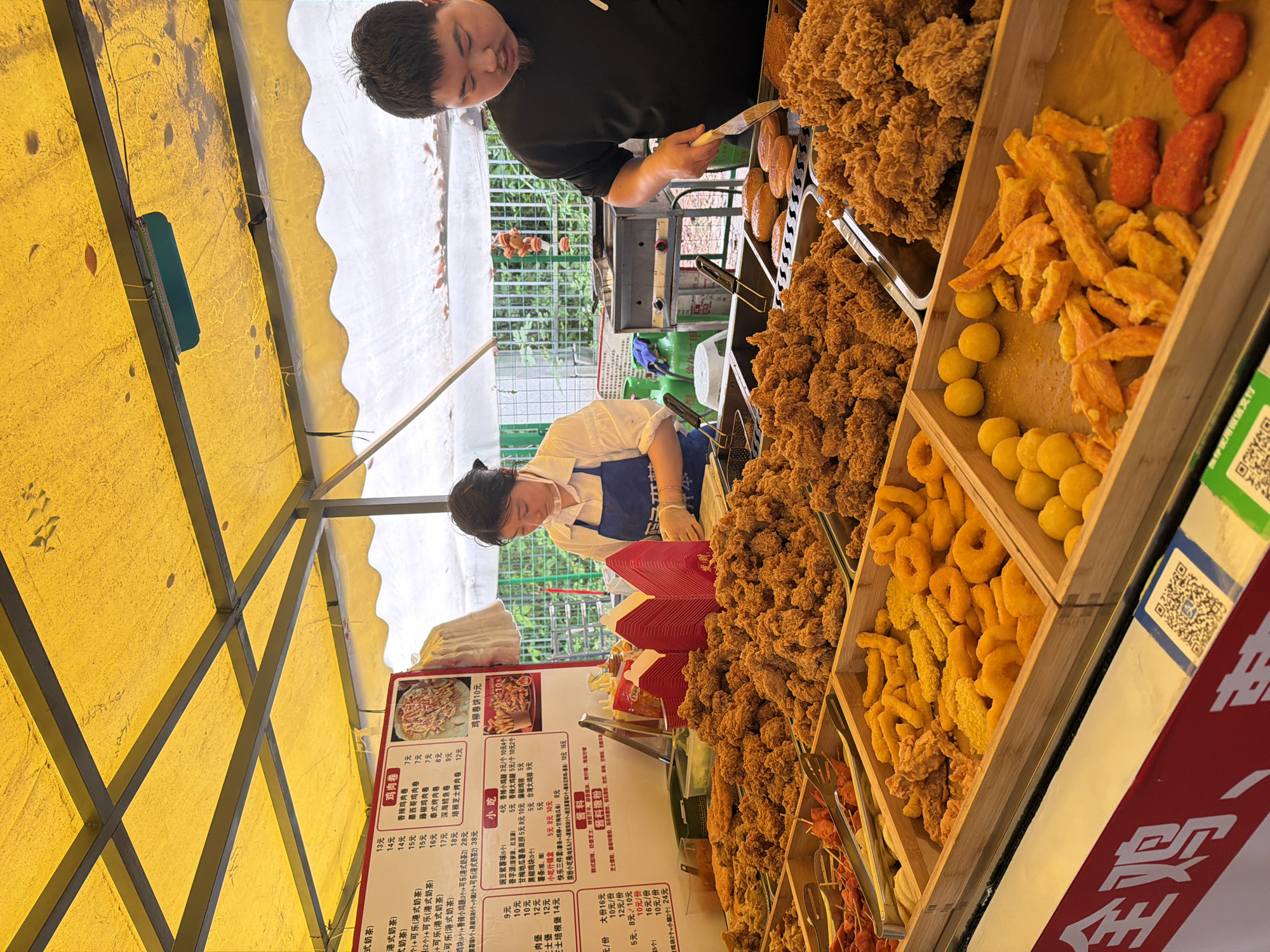 This image shows a food stall in Shapingba District, China, where two individuals are working. A man is using tongs to handle fried food, while a woman, wearing a white shirt and blue apron, is working at the counter. The stall is stocked with various fried foods, including chicken, sweet potato fries, donuts, and round balls. Menus are displayed on the side of the stall, detailing the food options and prices in Chinese. The stall is covered with a yellow awning.