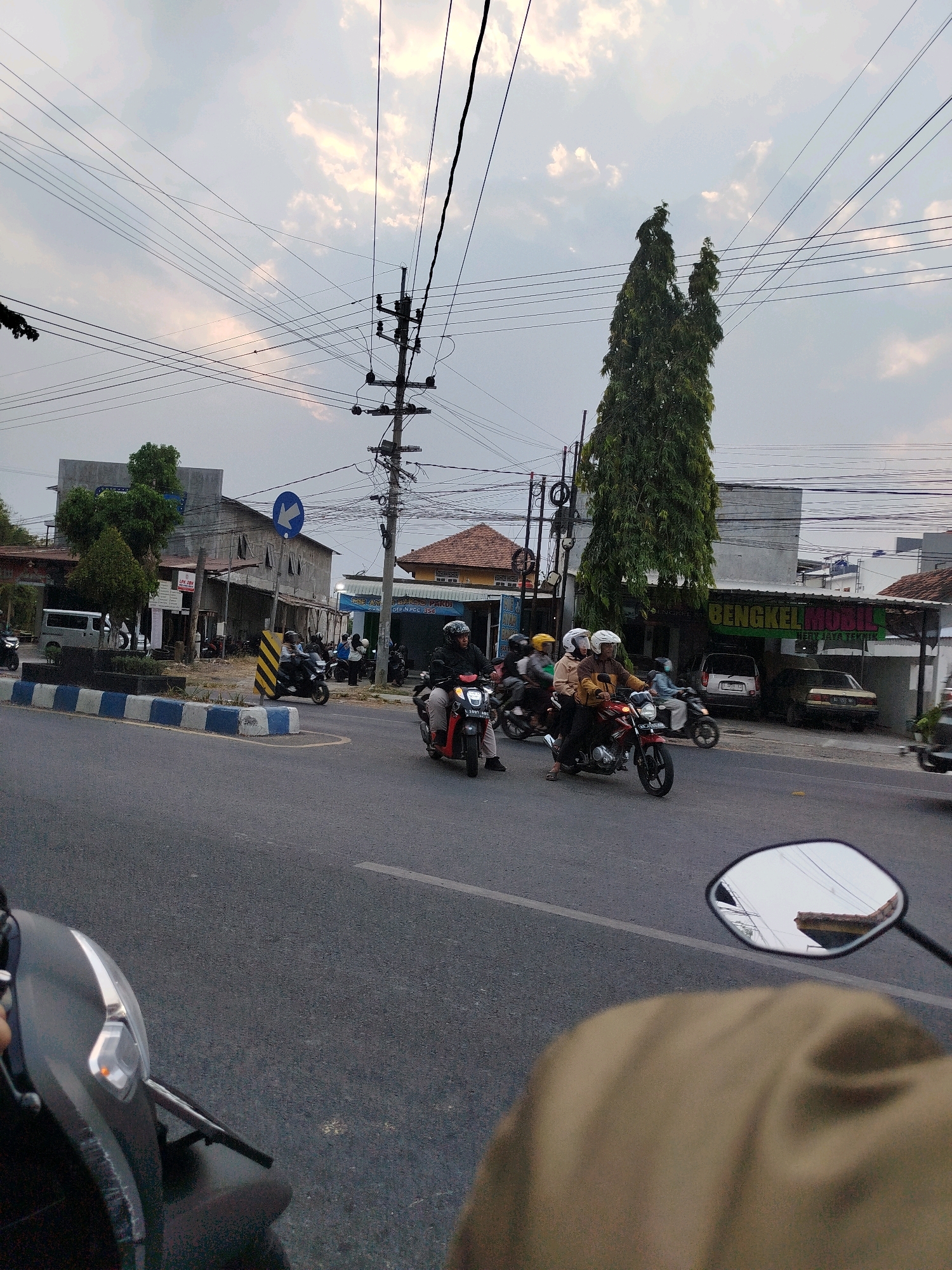 The image depicts a street scene in Cekok, Indonesia, with several motorcycles in motion. In the foreground, a person on a motorcycle is partially visible from a first-person perspective, their head and shoulder covered by brown fabric. The motorcycle's front headlight is illuminated. Further down the road, multiple motorcycles are traveling in the same direction. One motorcycle carries three people, all wearing helmets, and another has two people, also helmeted. A single rider is on a red motorcycle. On the side of the road, there are buildings, including what appears to be a car repair shop with a sign that reads "BENGKEL MOBIL". Utility poles with numerous overhead wires are prominent features of the urban landscape. The sky is overcast with some patches of lighter clouds. The road surface is asphalt, with a white line marking the lane. A blue and white striped barrier is visible on the left side of the road.