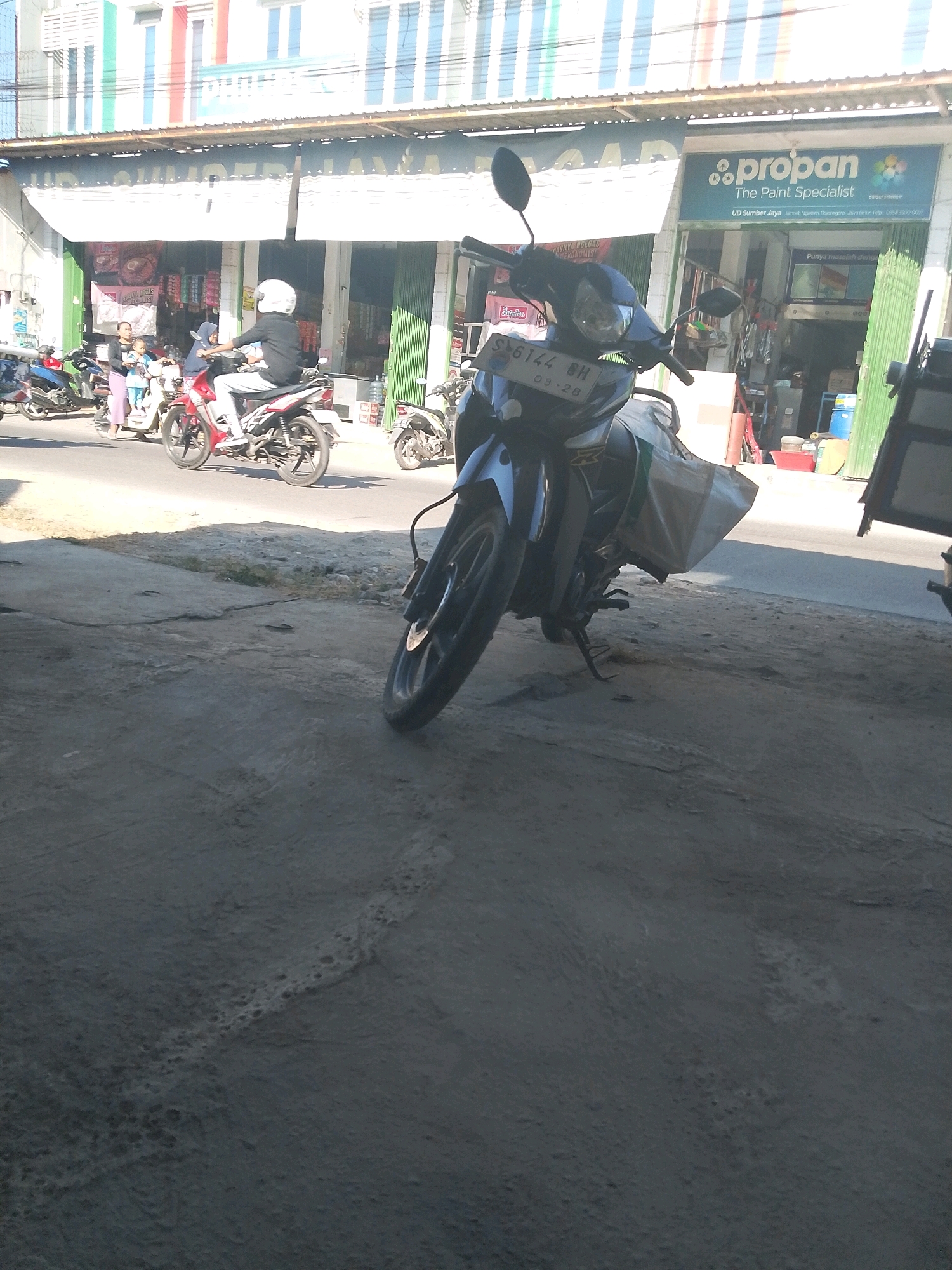 An eye-level, medium shot shows a street scene in Jampet, Indonesia, on a sunny day. In the foreground, a parked motorcycle is angled to the right, its front wheel and part of the engine visible. Behind it, on the paved street, several other motorcycles and a few people are visible. To the left, a woman in a pink shirt and a child in a light blue shirt are standing next to a white motorcycle. A person in a black shirt and helmet is riding a red and white motorcycle. Further back, a building with a blue awning and the text "Propan The Paint Specialist" is visible. The building has a green metal door and displays various paint-related products. The street is lined with shops and buildings, and the sky is clear and bright.