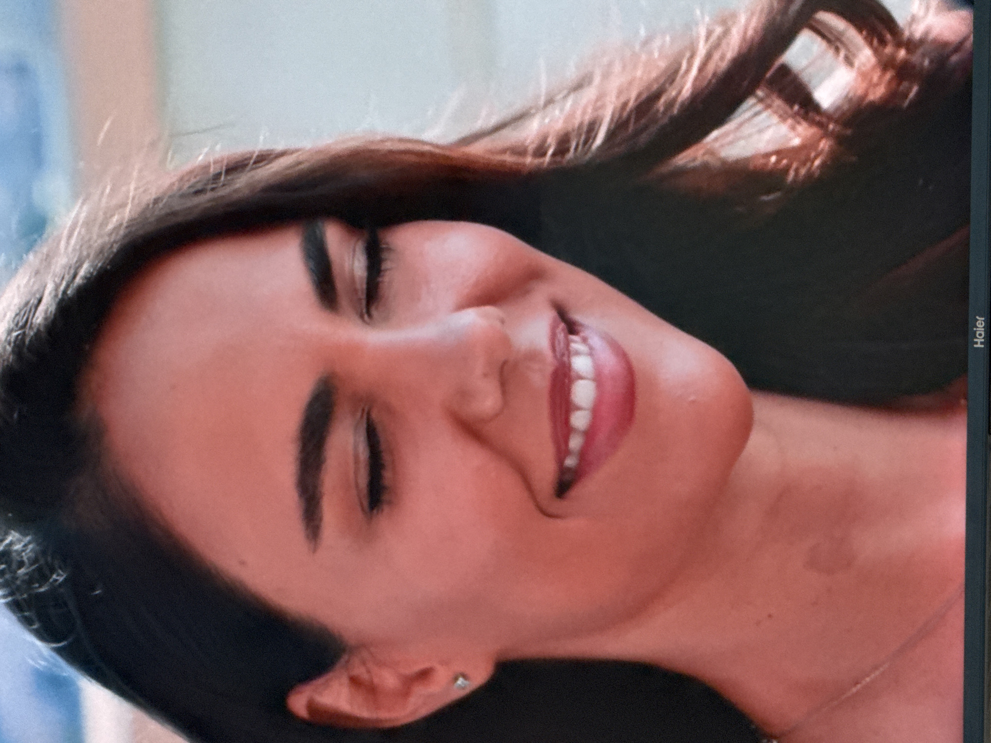 The image is a close-up, waist-up shot of a young woman with dark hair, smiling with her eyes closed. She is wearing a light-colored necklace and a small, silver stud earring in her left ear. The lighting is warm, casting a reddish hue on her skin. Her lips are parted, revealing white teeth, and she has well-defined eyebrows. The background is out of focus, appearing as a blurry wash of light blue and beige, possibly indicating an outdoor setting with natural light. There is a faint, darker mark on her neck. The bottom of the image shows a black bar with the word "Haier" in white text, indicating the image is displayed on a Haier monitor. The location of Uyutnoye, Ukraine, is not depicted or suggested by any elements within the image.