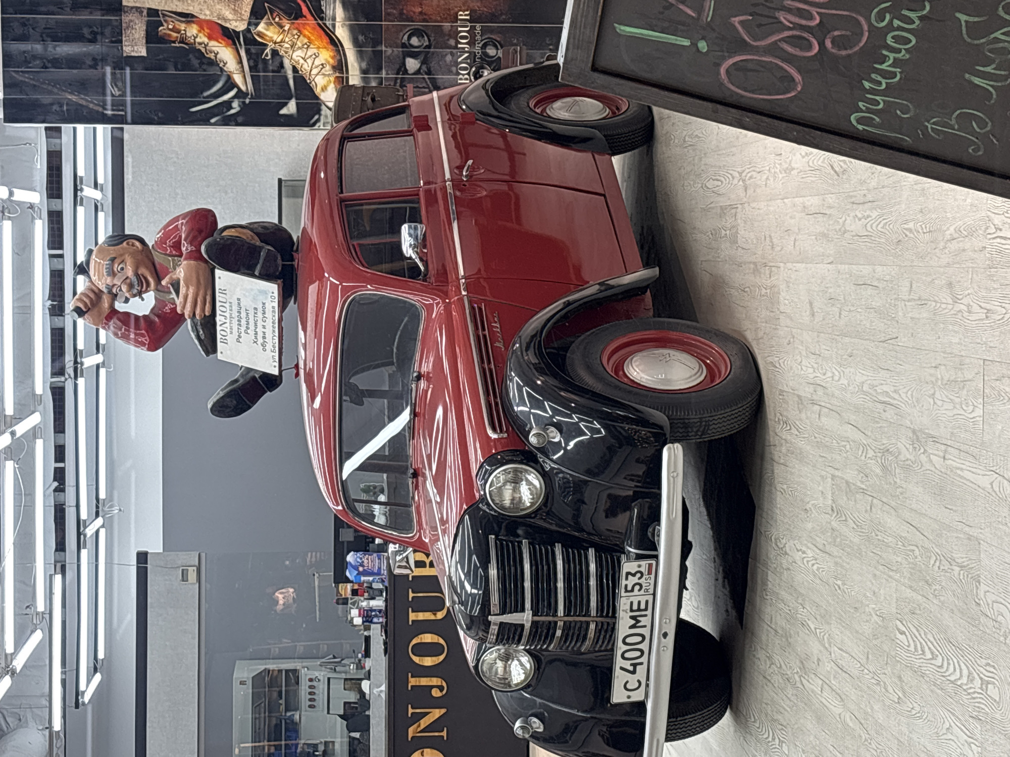 The image captures a quirky and somewhat unusual scene, likely inside a commercial space, possibly in Saint Petersburg, Russia, based on the Cyrillic text on signage and the license plate.
The central focus is a vintage red and black car, possibly a Moskvitch 400, which appears to be part of the decor or a display piece. It has a license plate that reads "C400 ME 53 RUS."
Perched on the side of the car is a figurine of a man with a distinctive mustache, dressed in a red shirt and dark pants. The figure holds a sign that reads "BONJOUR мастерская Реставрация Ремонт Химчистка обуви и сумок • ул. Бестужевская 10." This indicates the location is likely on or near Bestuzhevskaya Street, which is a street in Saint Petersburg.
To the right of the car, a black chalkboard with pink and green Cyrillic text leaning against it. The background includes glimpses of a retail or workshop environment, with shelves of items and bright overhead lighting. There's a poster on the wall depicting boots.