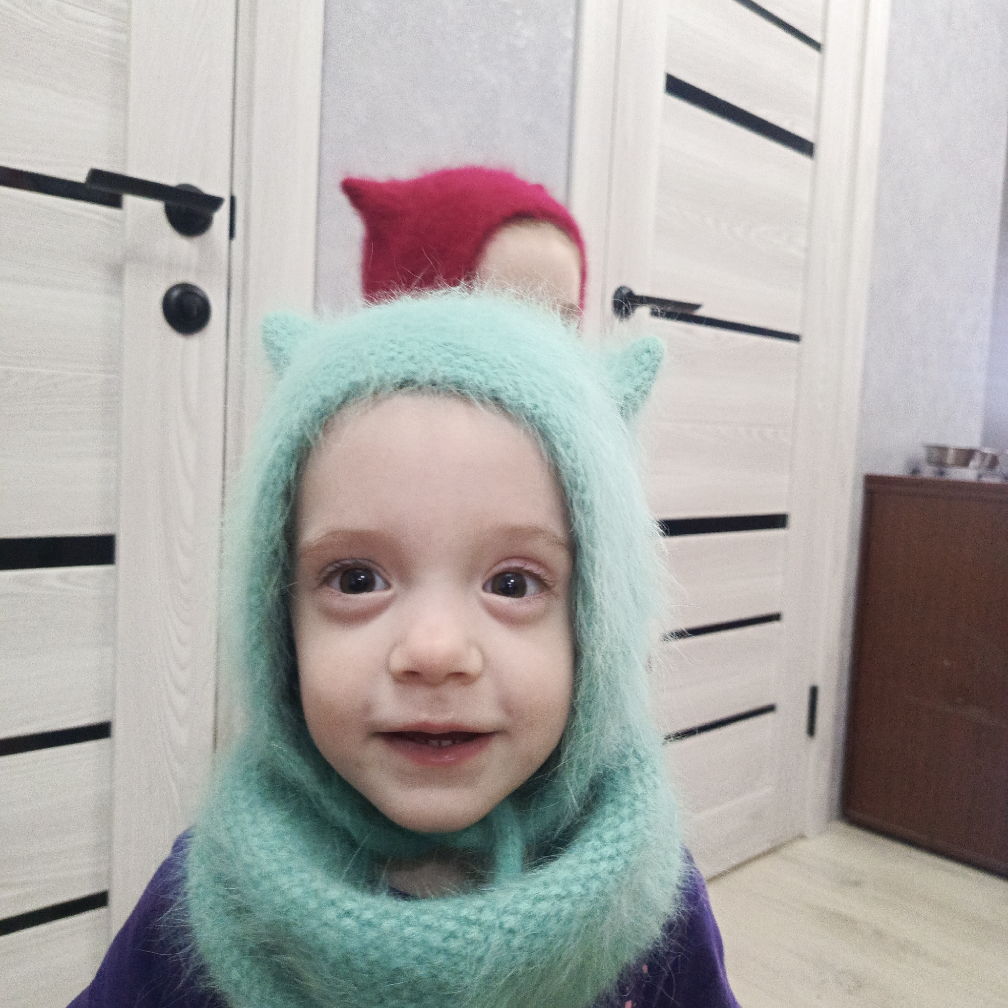 A young child wearing a turquoise fuzzy hooded scarf is smiling at the camera. Behind the child is another individual wearing a red hat with pointed ears. Two white paneled doors with black handles are partially visible in the background. A dark wood cabinet sits to the right, against a wall with patterned wallpaper. The image appears to be taken indoors. This image was taken in the city of Марковский, Russia.