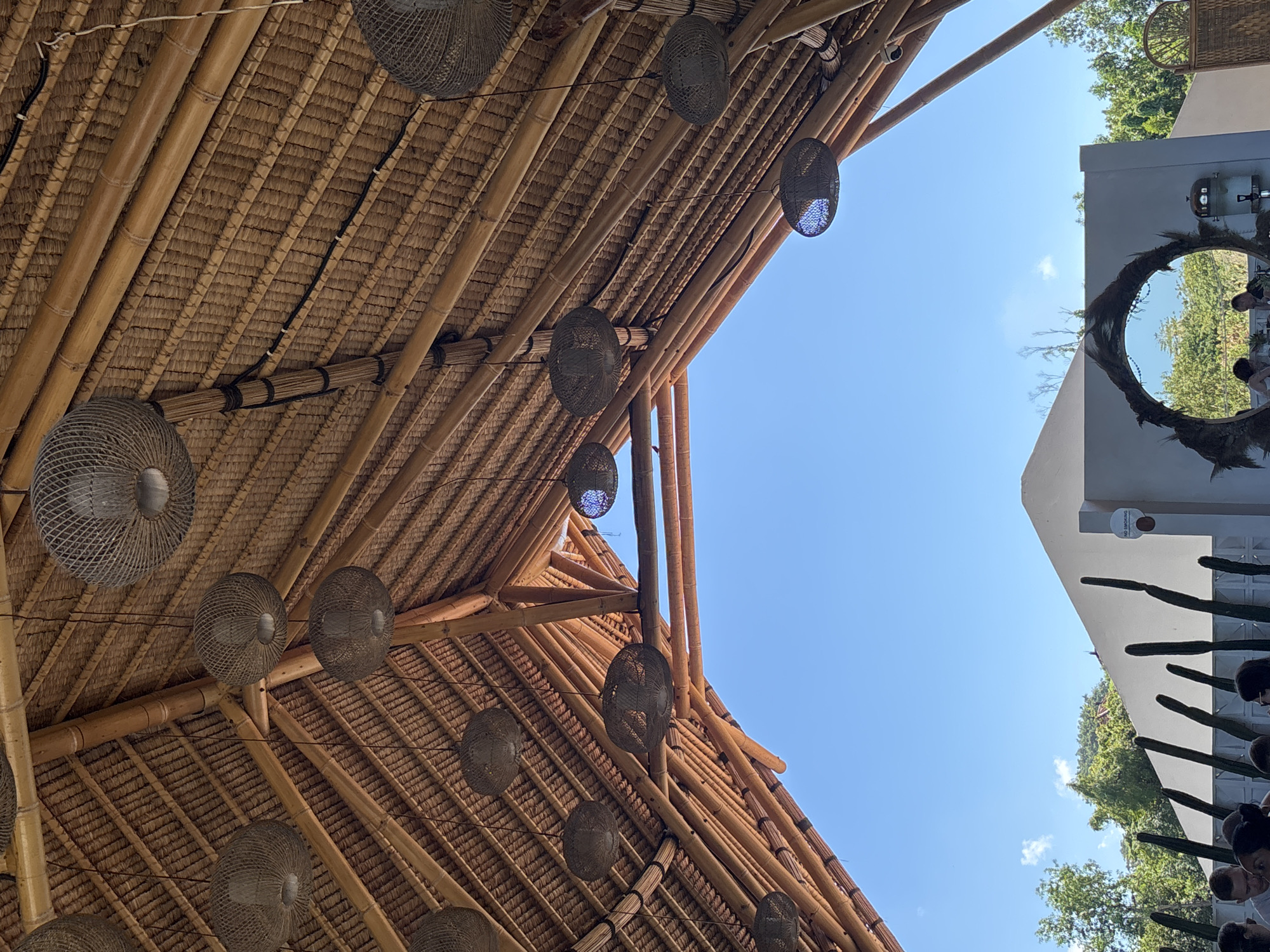 Captured from a low angle, the photograph features a thatch-roofed structure in Kuta Selatan, Indonesia, against a clear blue sky. The roof is constructed with bamboo supports and thatch, and a series of spherical woven light fixtures hang from the structure. To the right, part of a white building is visible, ornamented with tall cacti and a round decorative frame. The edges of people's heads are also visible at the bottom of the frame.