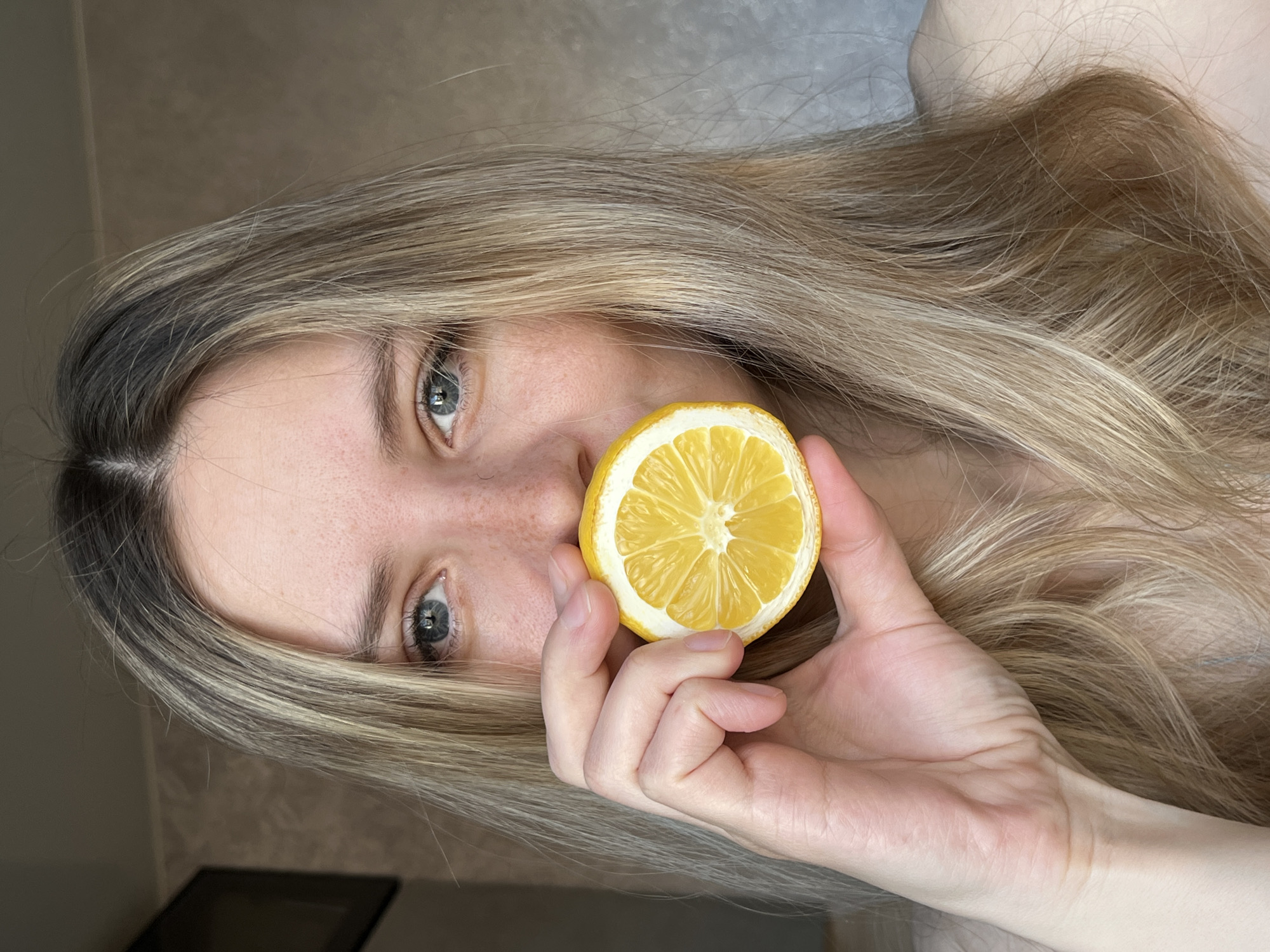 A person with fair skin and blonde hair holds a halved lemon up to their face, partially obscuring their nose. The background is a neutral-toned wall, with a dark object visible in the bottom left corner. The image appears to have been taken indoors. The city of Chaikovsky, Russia, cannot be determined from the visual information provided.