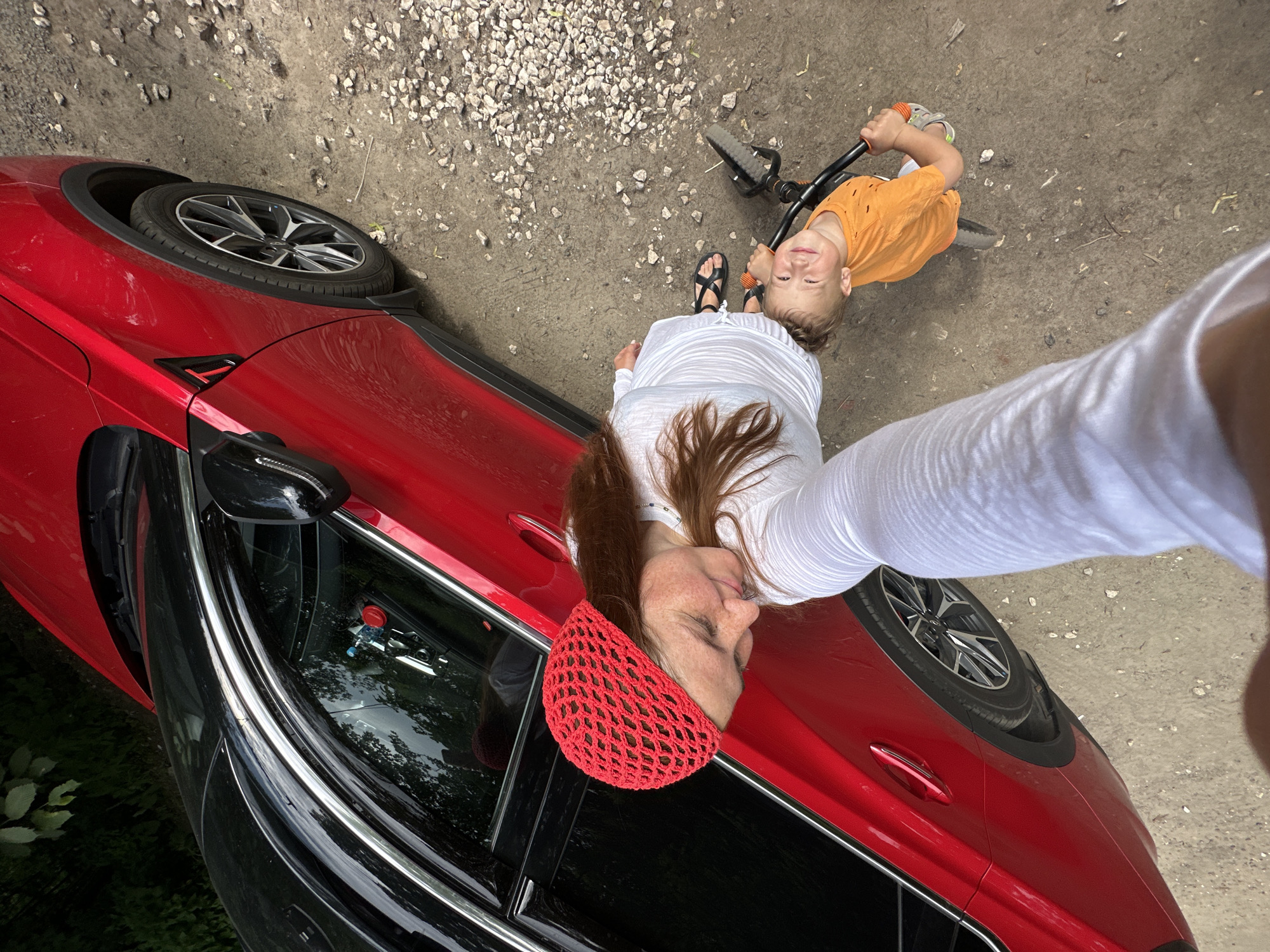 A woman wearing a red crocheted headscarf and a white top, with long auburn hair, is taking a selfie with her young son. The boy, wearing an orange t-shirt, is riding a small black tricycle. They are standing next to a red car. The ground is a mix of dirt and small white stones. In the background, there are trees and greenery. The image was taken in Nizhny Novgorod, Russia.