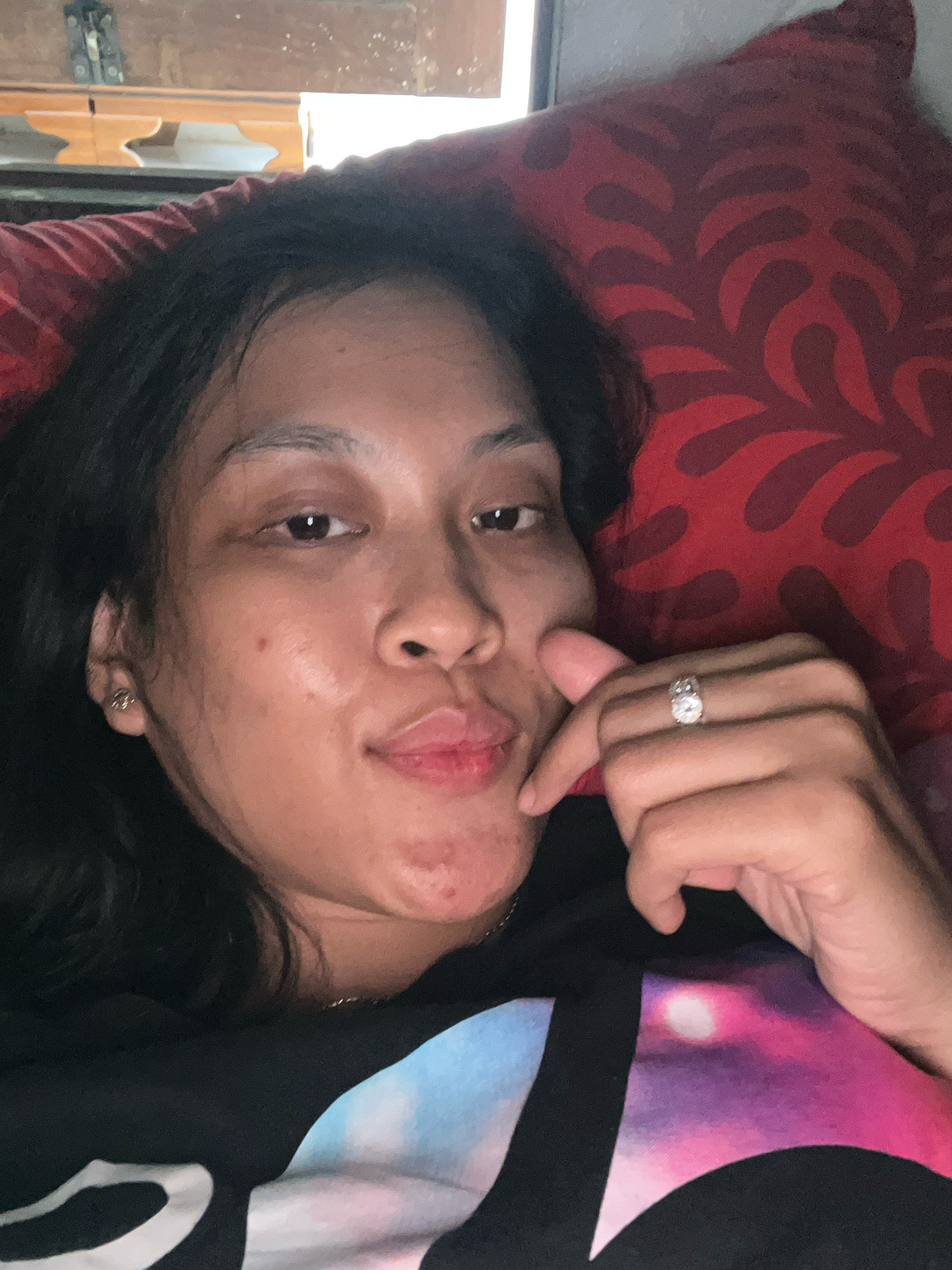 A close-up, high-angle, indoor selfie shows a person lying down and resting their head on a red patterned pillow. The person, who appears to be female, has dark hair, dark eyes, and light-to-medium skin tone. She is wearing a black t-shirt with a colorful galaxy-like print on the front. Her left hand is resting on her cheek, and she is wearing a large diamond ring on her left ring finger. The background suggests a room with wooden furniture visible, possibly in a home in Kecamatan Pati, Indonesia. The lighting is dim, and the focus is on the person's face and upper body.