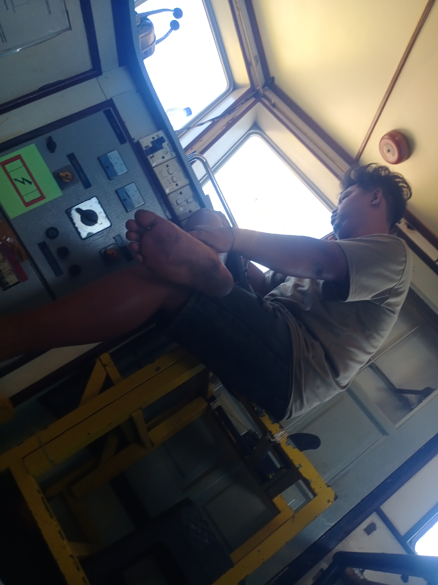 The image shows a man resting in what appears to be a control room, possibly on a boat or in a factory. The man, who is tanned, is wearing a light-colored t-shirt and dark pants. His left leg is bent, with his foot resting on his knee. He is looking to the right, away from the camera. The room is filled with various control panels and buttons, suggesting an industrial or operational setting. A yellow metal structure is visible in the foreground. The lighting indicates it is daytime, with natural light coming from a window. The location is identified as Siddo, Indonesia.