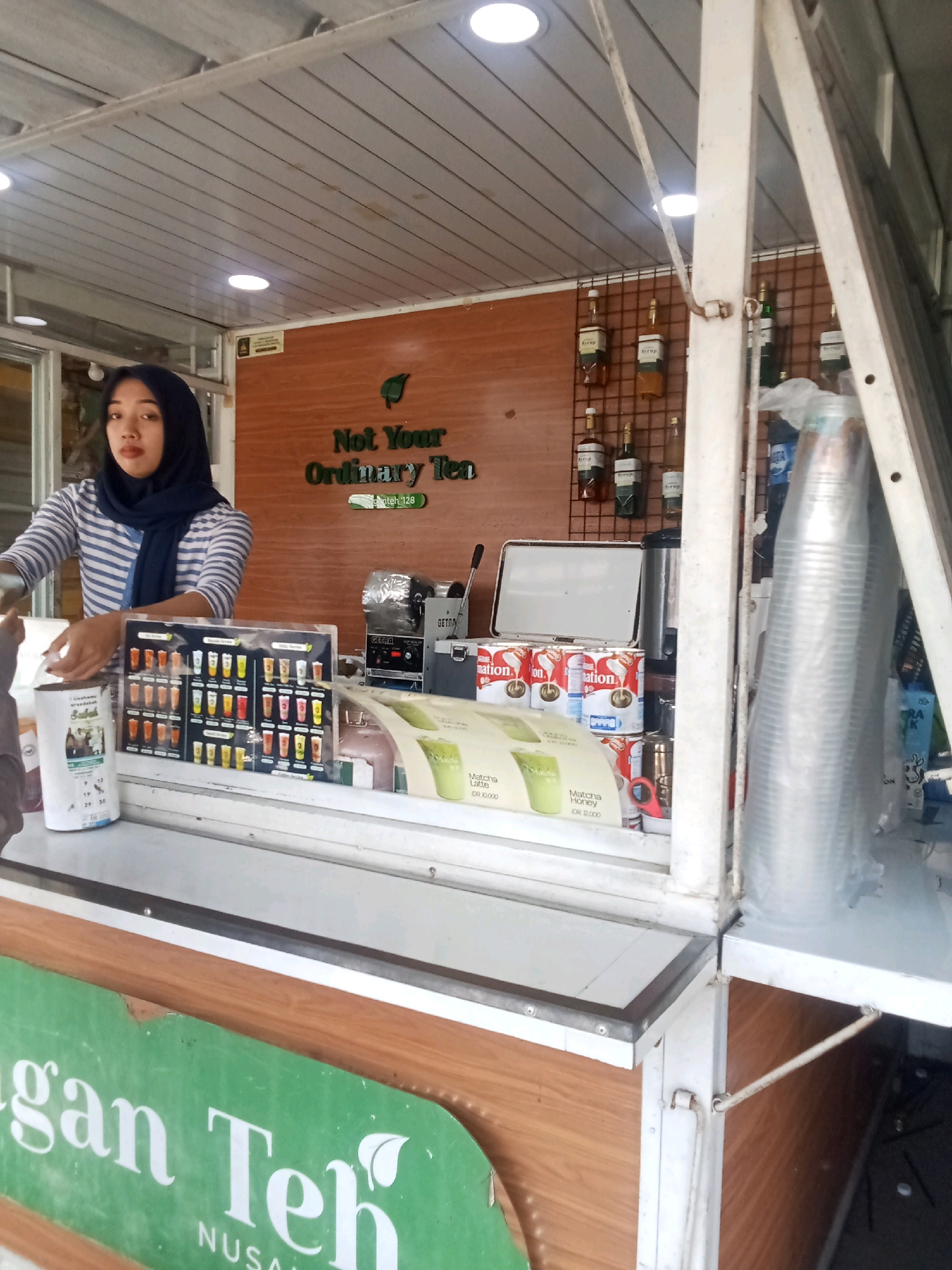 A young woman wearing a navy blue hijab and a striped long-sleeved shirt stands behind a counter in an outdoor food stall. The stall has a wooden facade with a green sign displaying white text that reads "gan Teh NUSAN" which is likely part of a larger company name related to tea. The woman is looking towards the camera with a neutral expression. In front of her, on the counter, is a menu board displaying various beverages with their prices. To the right of the menu are several cans of condensed milk and a thermal container. Behind the counter, shelves are stocked with bottles of syrup. The stall appears to be set up in a public area, possibly a street or market. The overall scene depicts a small business operation for selling tea and related drinks.