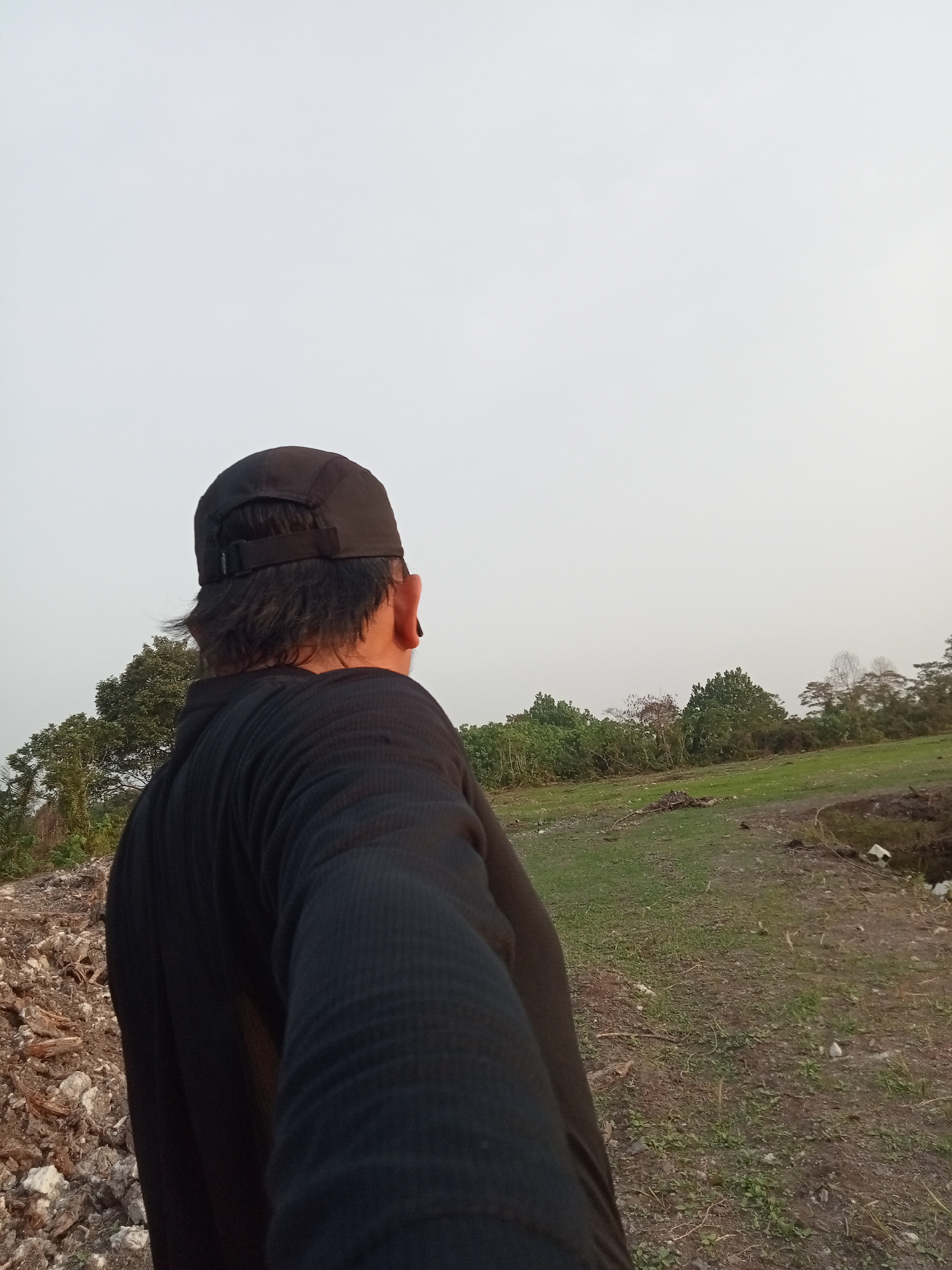 A person wearing a black long-sleeved shirt and a black baseball cap is standing outdoors, seen from behind and to the side, facing away from the camera. They are looking towards a landscape with a large expanse of sky. The ground in front of them is covered in dry grass and dirt. In the background, there is a line of trees and vegetation. The overall impression is of an open, natural setting, possibly in a rural area on the outskirts of Teluk Kenidai, Indonesia. The lighting suggests it could be late afternoon or early morning due to the soft, diffused light.