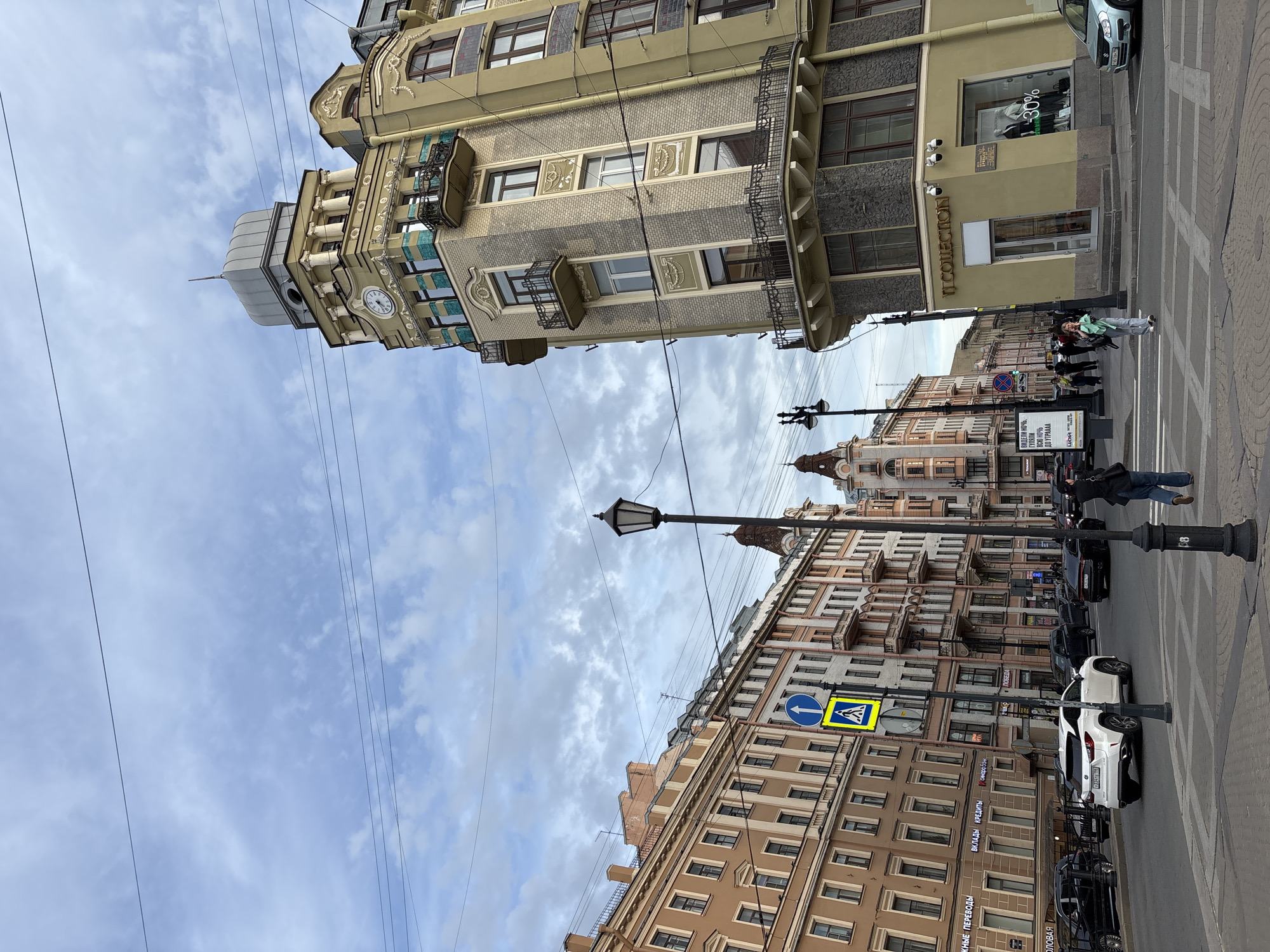 This image captures a vibrant street scene in Saint Petersburg, Russia, featuring grand architectural facades under an overcast sky. The primary subjects are the historic buildings lining both sides of the street, various vehicles, and pedestrians.
On the left, a particularly ornate, multi-story building stands out with a light-colored, possibly beige or pale yellow, facade adorned with intricate stucco details, decorative balconies, and a prominent dome-like structure on its roof, which includes a visible clock face. Its ground floor features shopfronts, one labeled "IT COLLECTION" and another displaying a "-30%" discount. Across the street, on the right, another row of multi-story buildings, primarily in warm brown and beige tones, presents a more traditional urban backdrop.
The street itself is visible with marked pedestrian crossings. Several cars are present, including a light blue Suzuki and a white Lexus, some parked along the curb. Pedestrians are seen on the sidewalks and crossing the street, engaging in typical daily activities. Overhead, utility wires crisscross the sky.
Visible details include a street lamp with the number "68" on its base and a pedestrian crossing sign (blue circular arrow and yellow diamond with a person walking). Text in Cyrillic is prominent on shop signs and an advertisement board. Notable text includes "ВКЛАДЫ КРЕДИТЫ" (Deposits Credits), "СМАРТФОНЫ" (Smartphones), "ПОБЕДА" (Victory), and an ad referencing Viktor Tsoi: "ВИДЕЛИ НОЧЬ, ВСЮ НОЧЬ ДО УТРАААА Цой" (We saw the night, all night until morning Tsoi).
The scene suggests a lively urban environment during daytime, characterized by a mix of historic architecture and modern city life, under a cloudy, bright sky.