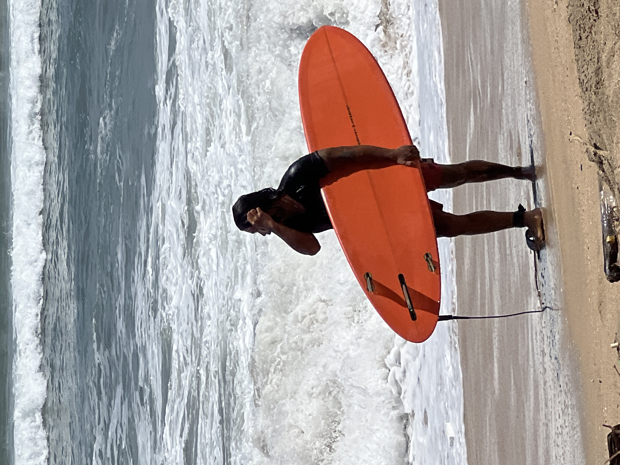 A man is standing on the shore at Kuta Beach, Indonesia, holding an orange surfboard. He is wearing a black wetsuit top and red shorts. The man is looking out at the ocean, and his hand is raised to his forehead as if shielding his eyes from the sun. Waves are crashing on the beach behind him. The sand is wet and appears to be packed down by the water. Some debris, including a plastic bottle and pieces of wood, are visible on the sand. The water is a murky blue-green, and the waves are white with foam. The sky is not visible in the frame.