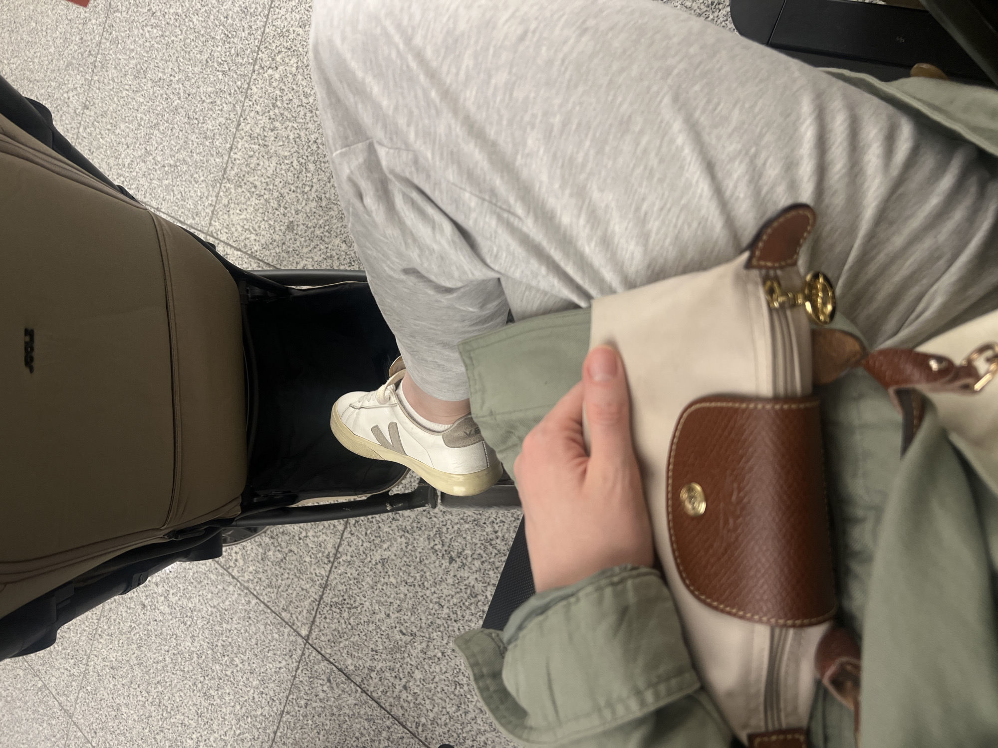 The image shows a first-person perspective of someone seated, likely at an airport based on the tiled flooring. The individual is wearing grey sweatpants, a green jacket, and white sneakers with a light-colored heel patch. They are holding a Longchamp-style handbag with a brown leather flap and gold hardware. To the left, a beige stroller is visible. The location of Khimki, Russia, is not verifiable from the provided image.
