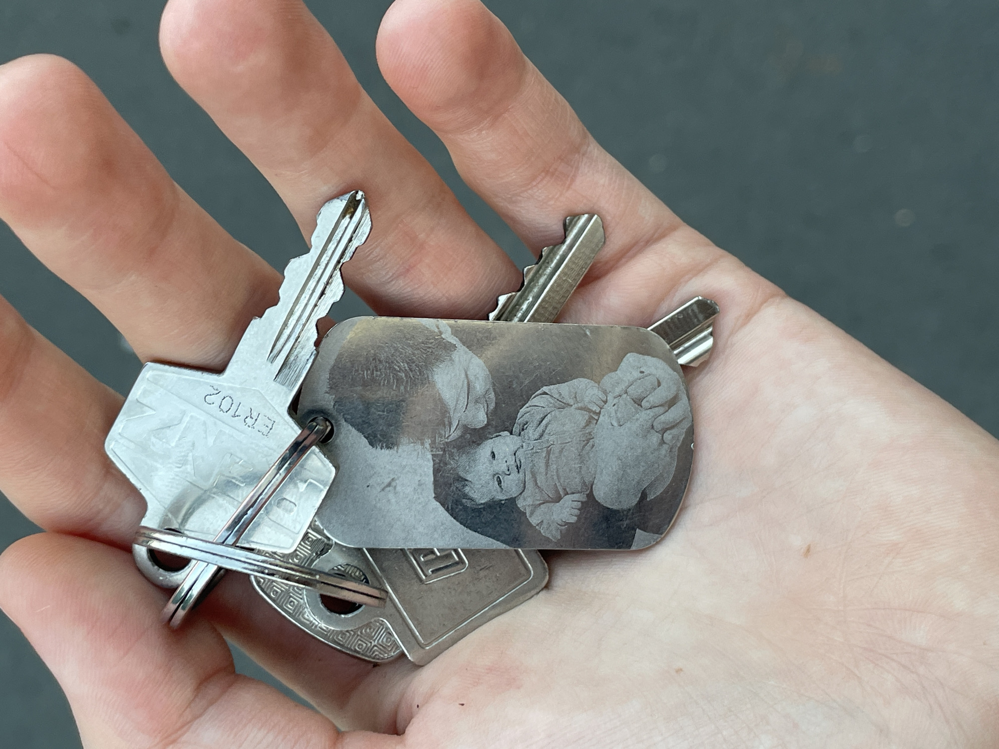 A person's hand is holding a set of keys. Among the keys is a rectangular keychain with an engraved image of a baby being held by an adult. The image is monochrome and appears to be a photograph transferred to metal. The keys are silver-colored and appear to be standard house or car keys. The background is out of focus and shows a dark gray, asphalt-like surface, suggesting an outdoor urban environment. The lighting is bright, indicating daytime. The specific location within Nidda, Germany, is not discernible from the image itself.