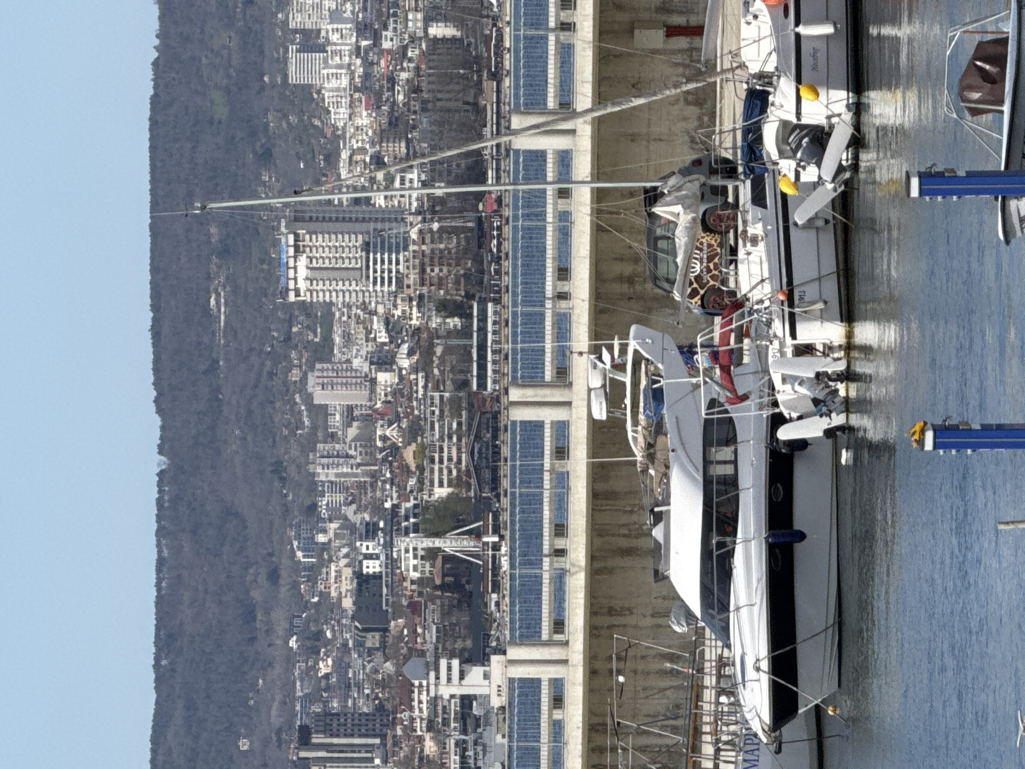 This vertical image captures a marina scene in Varna, Bulgaria, featuring boats docked in the foreground, a sprawling city in the midground, and forested hills in the background, all under a clear blue sky. The scene suggests a bright, sunny day.
In the immediate foreground, two boats are moored in calm, reflective water. The boat on the left is a white sailboat with a dark blue stripe, displaying the name "MackroN" on its stern and "Yamaha" on an outboard motor. It has a red life buoy and a notable giraffe-print cover over a window or hatch. Adjacent to it, on the right, is a white motor yacht with a dark hull stripe, and partial text "MAP..." (possibly "MAPLE") is visible on its stern.
Behind the boats, a concrete dock or pier structure runs horizontally, adorned with multiple large, blue, rectangular panels, likely solar panels or reflective window arrays. Beyond this structure lies the dense urban landscape of Varna, characterized by numerous buildings of various sizes and architectural styles, extending across the midground.
The background is dominated by a long, dark, densely forested hill or mountain range, stretching across the horizon. The sky above is a uniform, pale blue, devoid of clouds. No specific human activity is discernable, but the boats are clearly moored in a harbor setting.