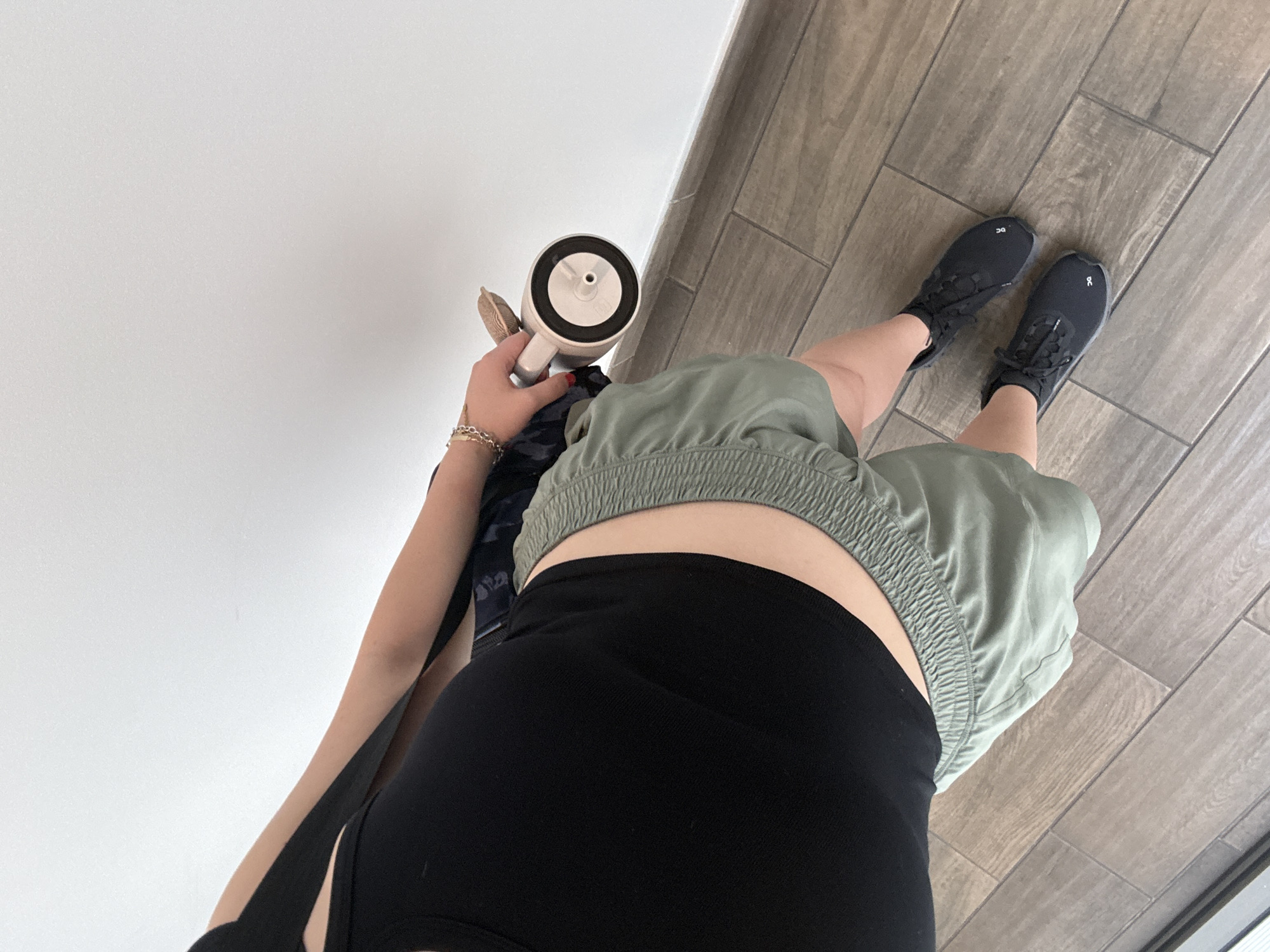 An overhead shot captures a person standing indoors in Mexico City, Mexico. The individual is partially visible, showing their torso, arms, and legs. They are wearing a black sports bra or crop top, olive green elasticized shorts, and black athletic shoes. In their left hand, they hold a light-colored tumbler or water bottle with a black lid and a central straw opening. Several gold-colored chain bracelets adorn their left wrist. The black athletic shoes have a distinct white "OC" logo visible on the top of each shoe. The background features a plain white wall on the left side of the frame and light brown, wood-look rectangular floor tiles covering the ground. The lighting suggests it is daytime. The person appears to be casually posing or standing, showcasing their comfortable, active attire.