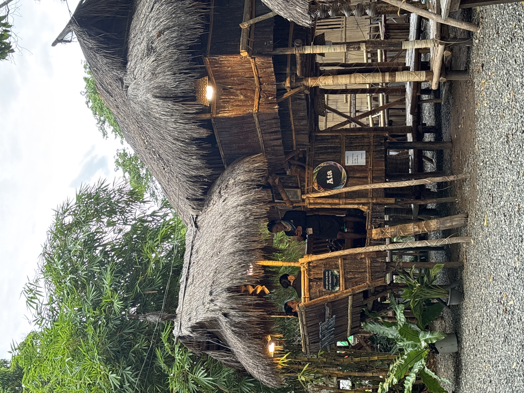 The image shows a rustic, stilted structure made of bamboo and thatch, resembling a traditional Filipino hut. It is located outdoors, likely in a tropical setting, with lush green bamboo trees and foliage surrounding it. The structure has multiple levels and open-air platforms, with thatched roofs. Several people are visible. On an upper level, a person appears to be serving or interacting with two seated individuals. Another person, dressed in what looks like a uniform, stands on a lower platform, holding a smartphone. A circular sign with the word "Ata" and some decorative elements is affixed to the structure, along with a smaller sign with text in Filipino. The ground is covered in gravel. This scene is evocative of a unique dining or cultural establishment, possibly found in Davao City, Philippines, given the context of its typical construction and vegetation.