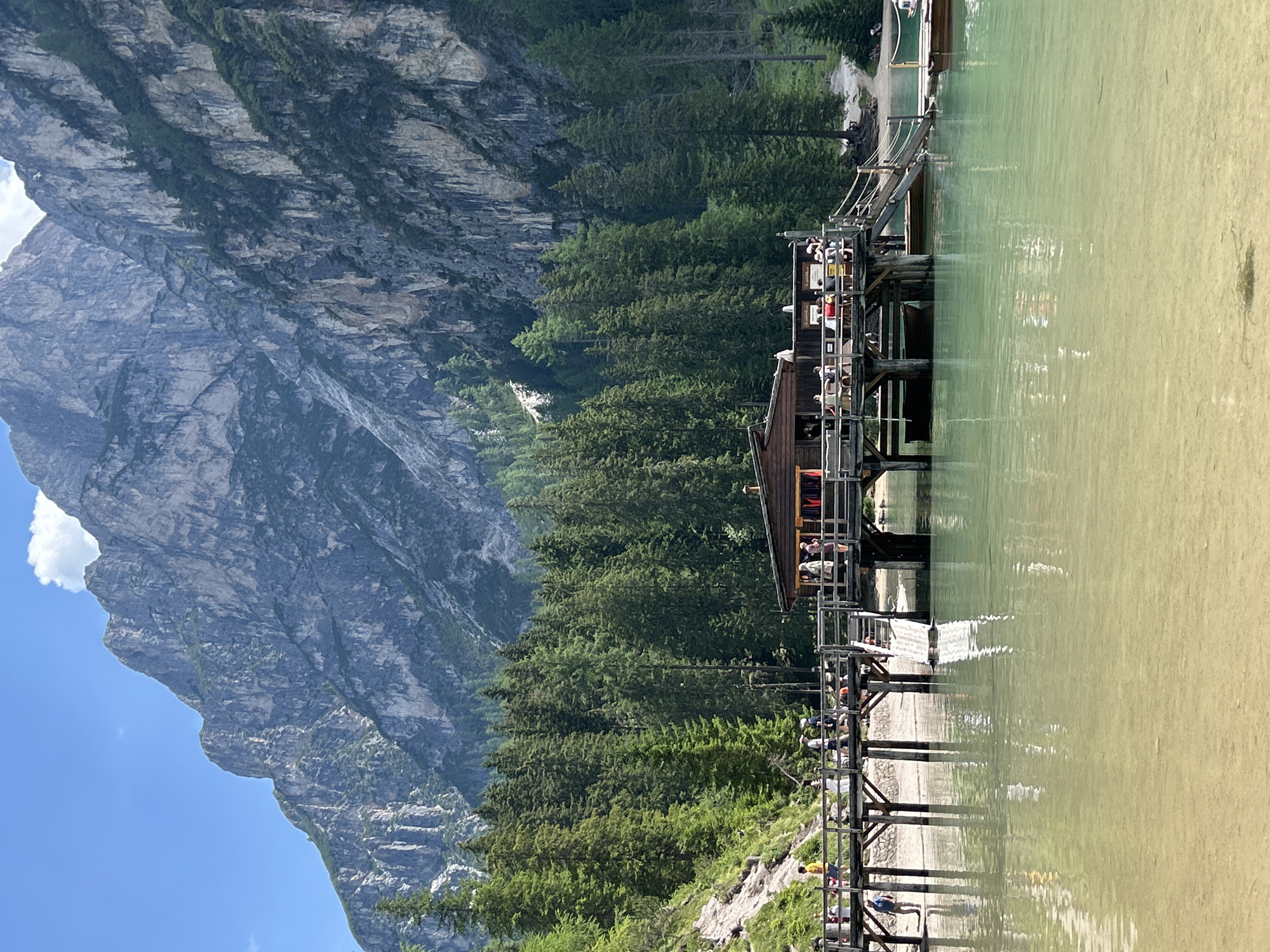 A scenic view of a lake with a wooden pier, dense green trees, and a large, rugged mountain in the background. The lake water is a clear, pale green, with sunlight glinting off its surface and showing the sandy bottom near the shore. The wooden pier extends into the lake and is populated with numerous people, some standing and some sitting. A small wooden structure is situated on the pier. The mountain is imposing, with its rocky, textured surface and patches of green vegetation. A clear blue sky with a few white clouds is visible above the mountain. The scene suggests a bright, sunny day, likely during warmer months, indicating a popular tourist or recreational area. The people on the pier appear to be enjoying the scenery, possibly taking photos or simply relaxing. The overall atmosphere is one of natural beauty and leisure.