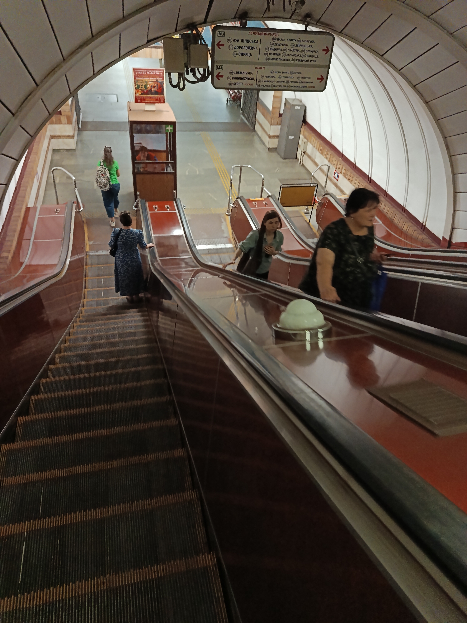 The image shows an underground metro station with a woman in a blue dress riding an escalator downwards. She is in the foreground of the image, and the escalator extends below her. To her left, another escalator is visible, also descending, with two women riding it. One woman is wearing a green shirt and jeans, and the other is wearing a black patterned shirt. In the background, there are signs indicating metro line information and directions, as well as a ticket booth. The station has a large arched ceiling.